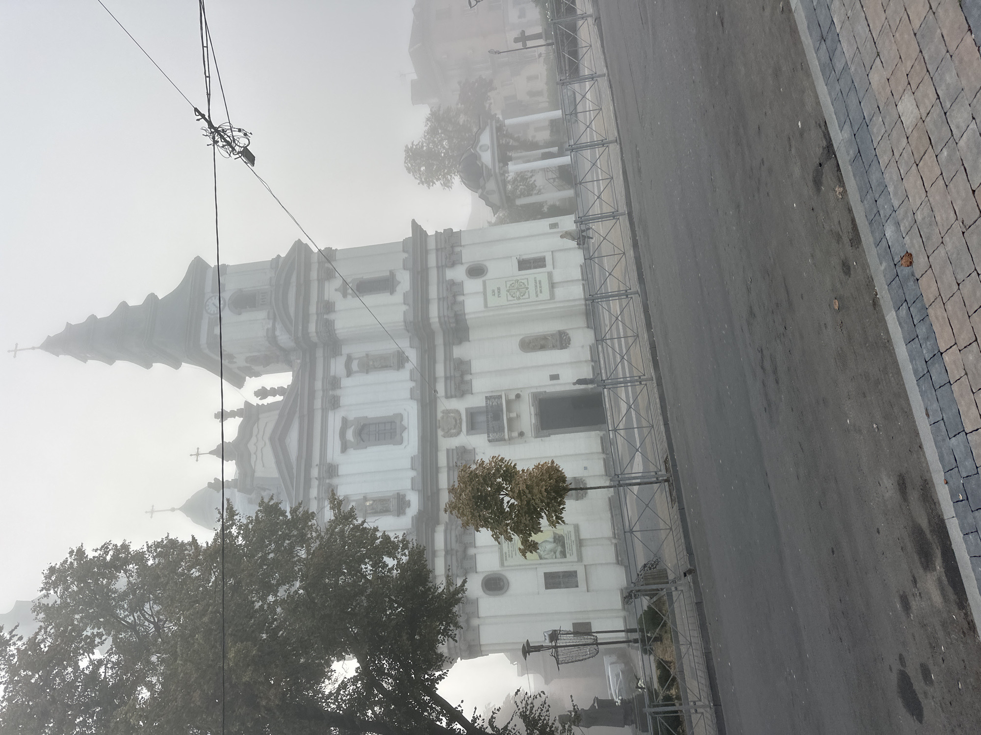 A church with white facades and a tall spire is partially obscured by fog in Ternopil, Ukraine. The church features Baroque architectural elements, including columns, ornate carvings, and a prominent clock face on its tower. A small tree stands in front of the church, and a statue is visible to the left. A metal fence separates the church grounds from the street, which is paved with asphalt and bordered by a sidewalk with patterned pavers. Overhead, power lines and a tangled mass of wires are visible. The overall atmosphere is hazy and subdued due to the fog.