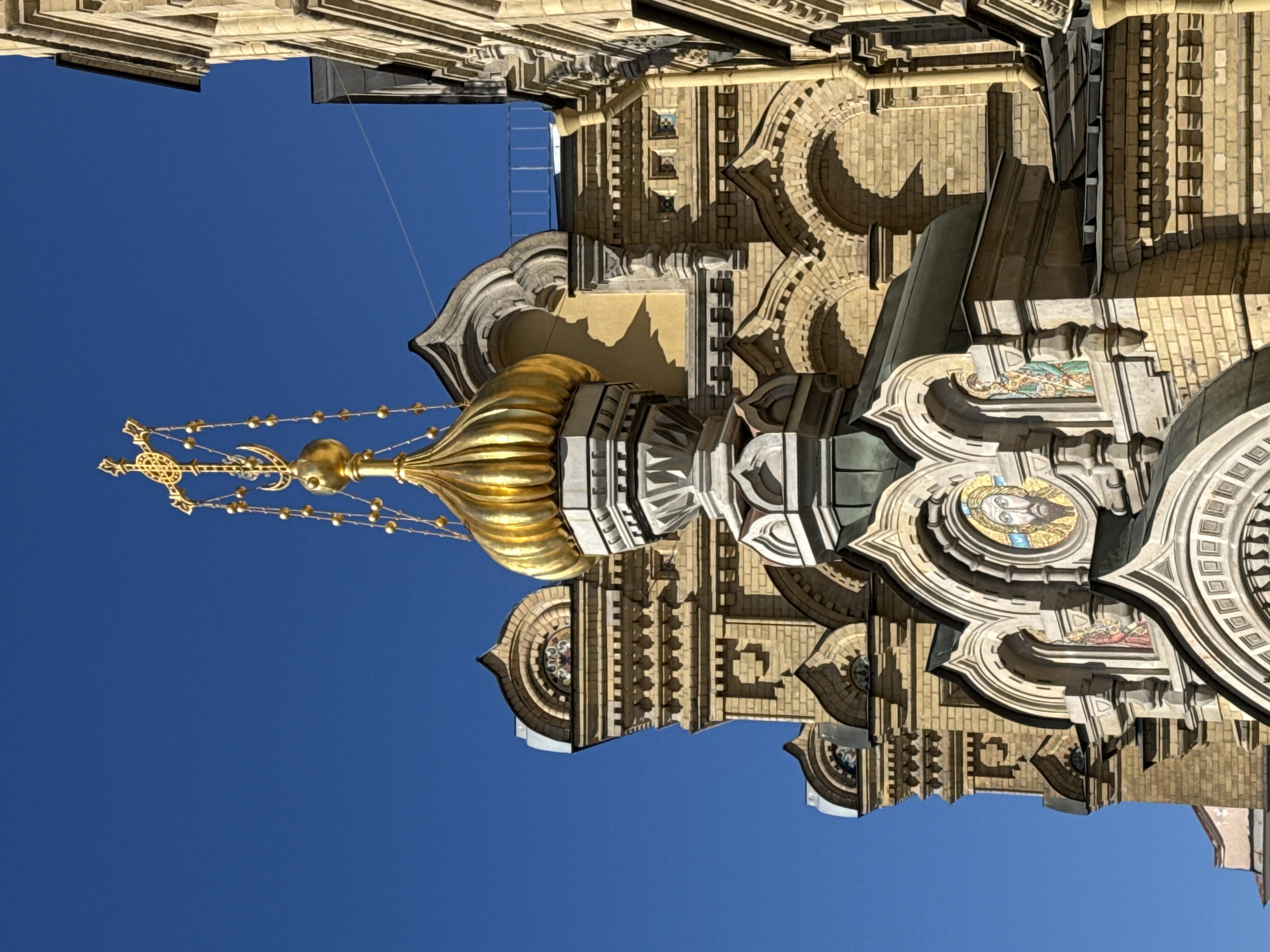 The image depicts the Church of the Savior on Spilled Blood in Saint Petersburg, Russia. The architectural details showcase the church's onion dome topped with a golden cross against a clear blue sky. Elaborate brickwork, ornate mosaics, and decorative elements characterize the building's facade.