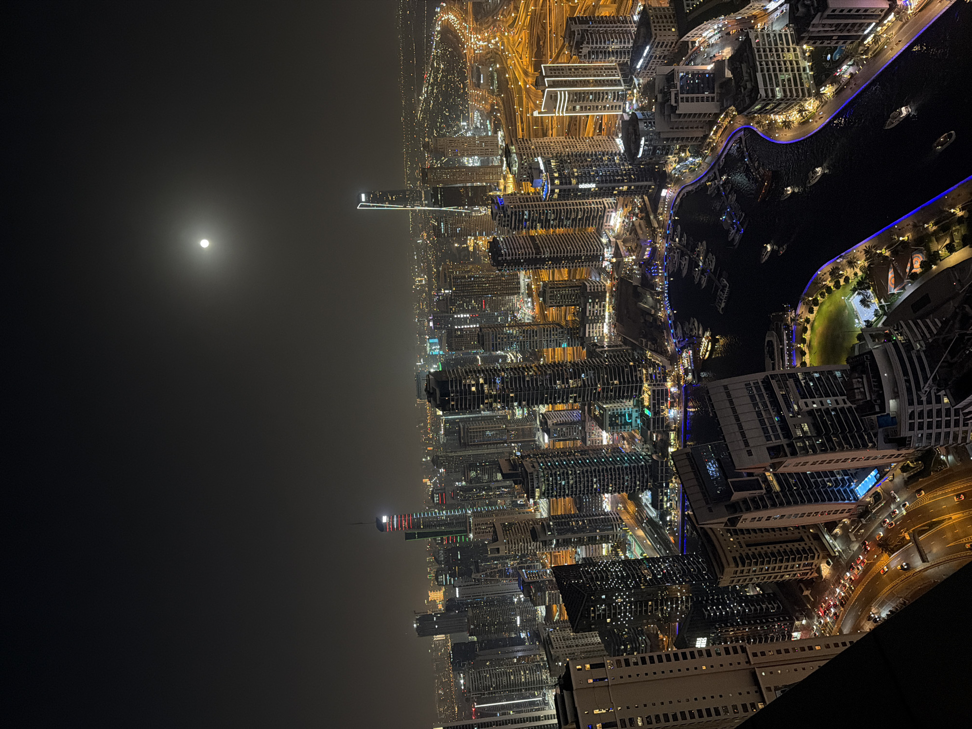 An aerial, nighttime view looking towards the city of Dubai, United Arab Emirates. The moon is visible in the sky. The city is lit with lights from buildings and roadways. A body of water curves through the city, with boats visible. Tall buildings are densely clustered in the foreground and stretch into the distance. A building edge is visible in the bottom right corner.