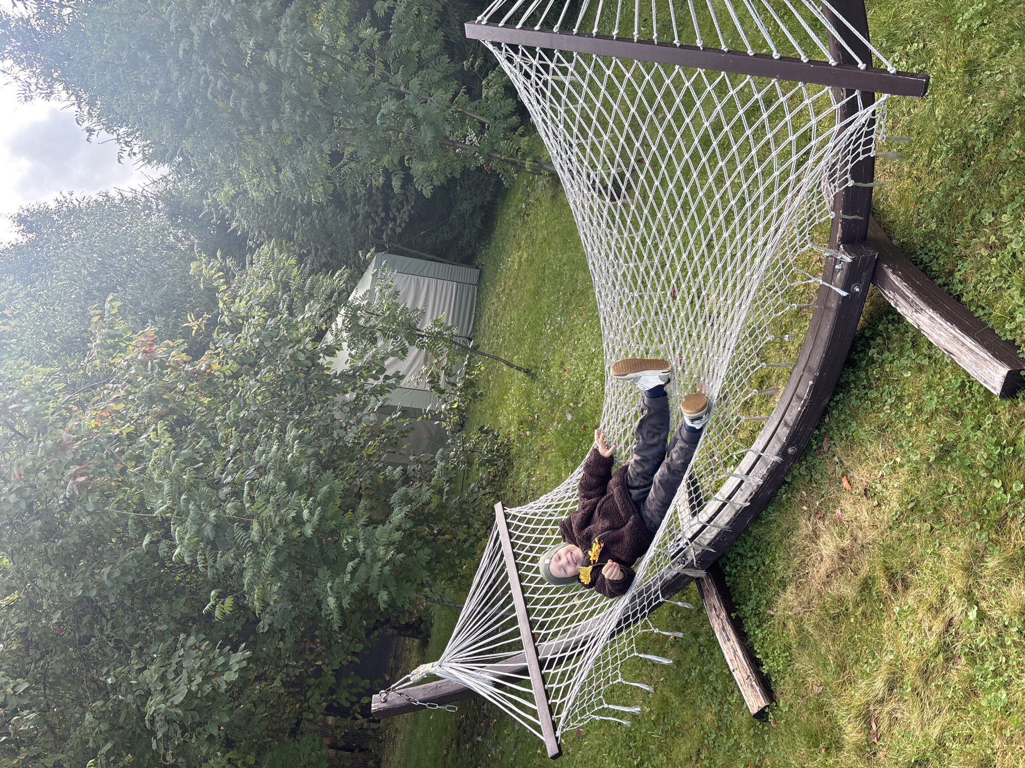 A young person is lying in a white rope hammock in a yard, with trees and a shed in the background. The person is wearing a green beanie, a brown fuzzy jacket, and camouflage pants. They are holding two yellow leaves and smiling at the camera. The hammock is suspended from a wooden frame, which rests on a grassy lawn. The shed is light gray and appears to be made of corrugated metal. The trees are dense and green, suggesting a temperate climate. The scene is set in the village of Rozhdestveno, Russia.