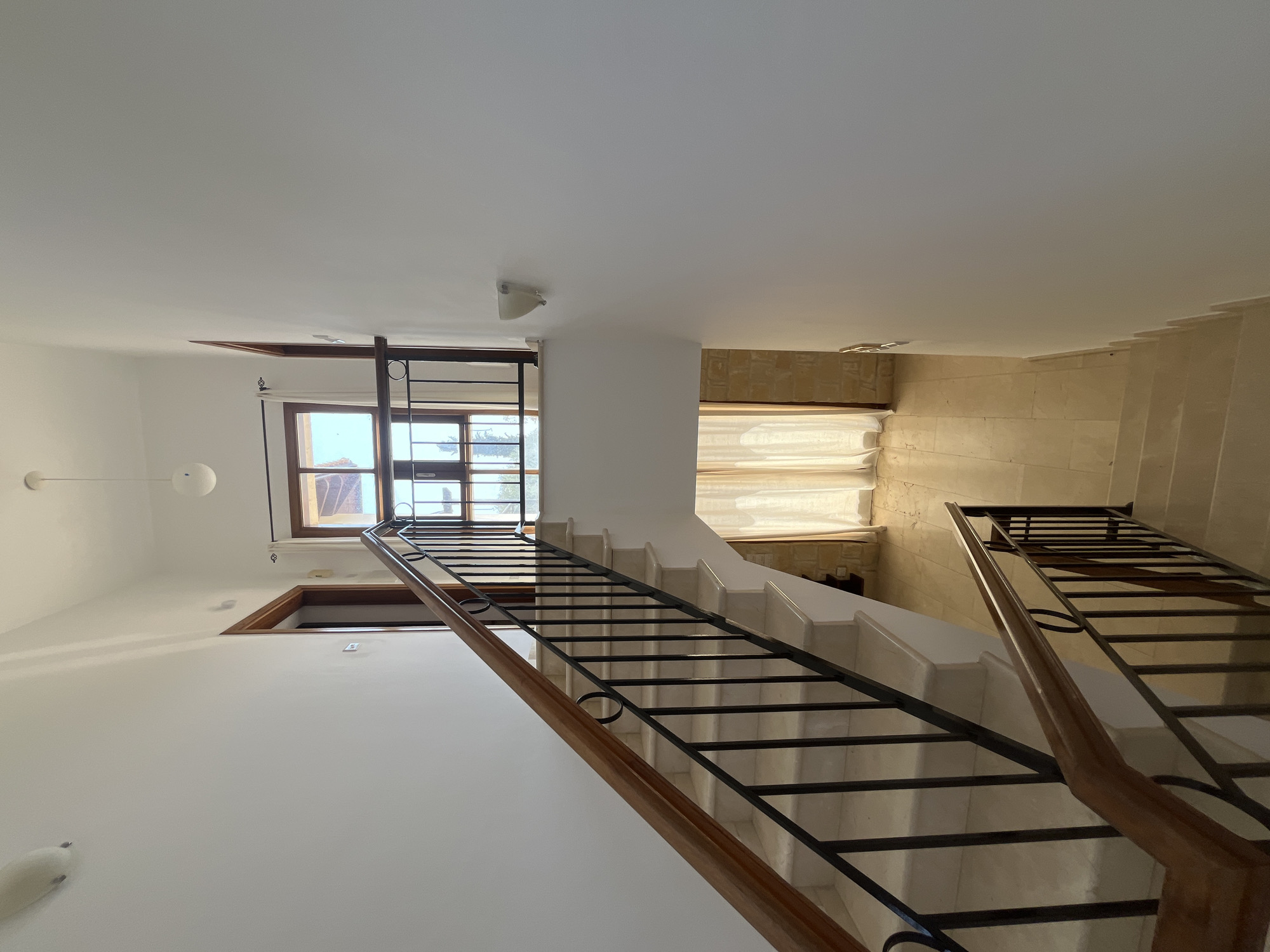 The image shows an interior staircase in a residential building, likely in Aphrodite Hills, Cyprus. The staircase is made of light-colored marble with a wooden handrail and black wrought-iron balusters. It ascends from a tiled floor, where sheer cream curtains hang over a window. At the top of the stairs, there is a landing with another window and a door. The walls are painted white, and there are two wall-mounted light fixtures. The overall impression is one of a clean, well-lit, and modern interior.