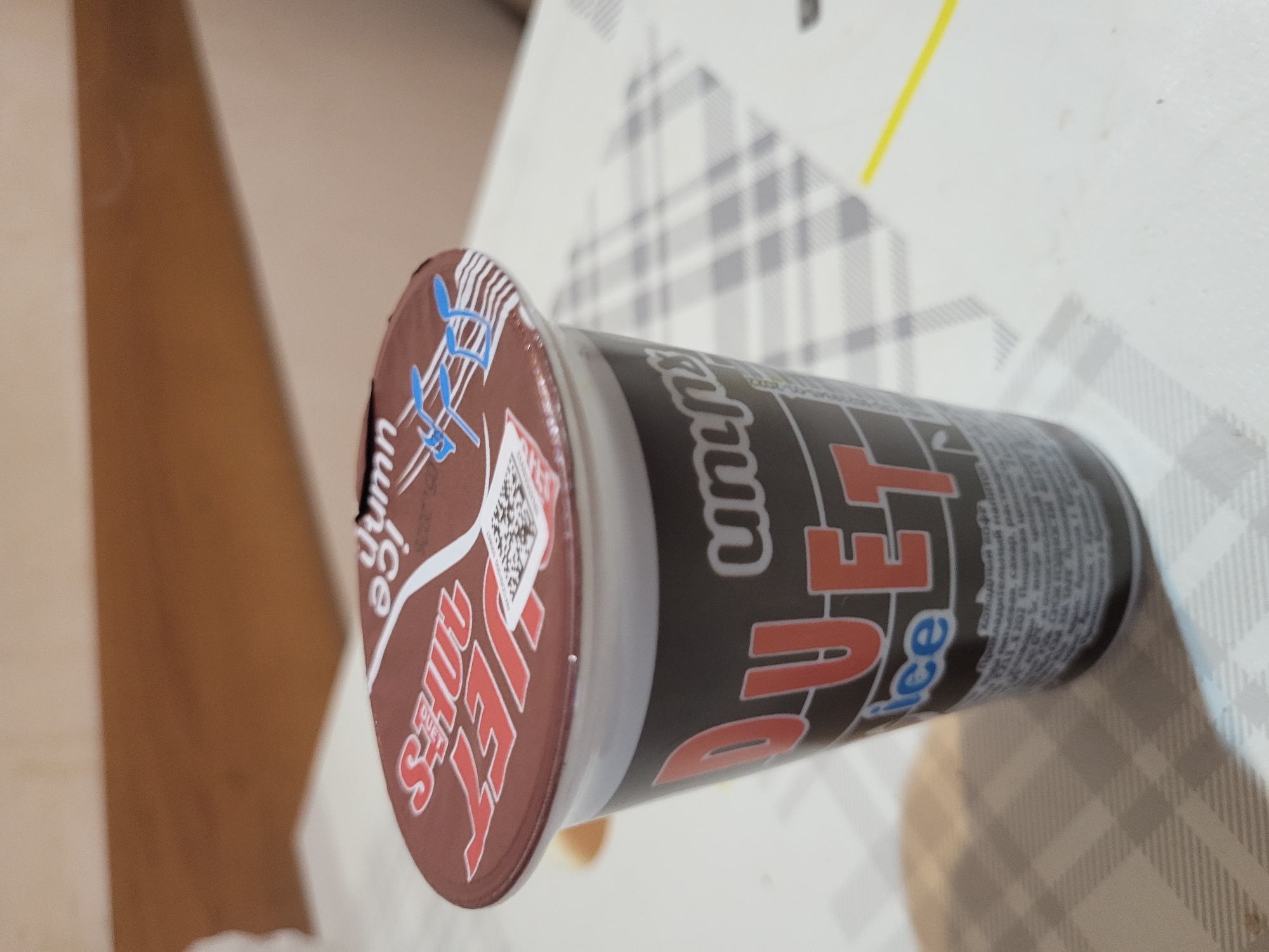 The image displays a cup of ice cream, likely chocolate-flavored, positioned horizontally on a table. The cup features a brown lid with a white rim and what appears to be a stylized blue and white musical note or design, alongside partially visible text like "Shuu" and "ice cream," and a QR code. The main body of the cup is dark gray with prominent orange and white text spelling "JET Duet" and "ice." The background consists of a white and gray checkered tablecloth with a distinct yellow line running across it, suggesting an indoor setting. Further in the background, a plain brown wall or surface is visible. There are no people or specific activities occurring in the frame; the image focuses solely on the product. The time of day, weather, and emotions are not discernible. While the location context of Yerevan, Armenia is provided, there are no explicit visual cues within the image to confirm this.