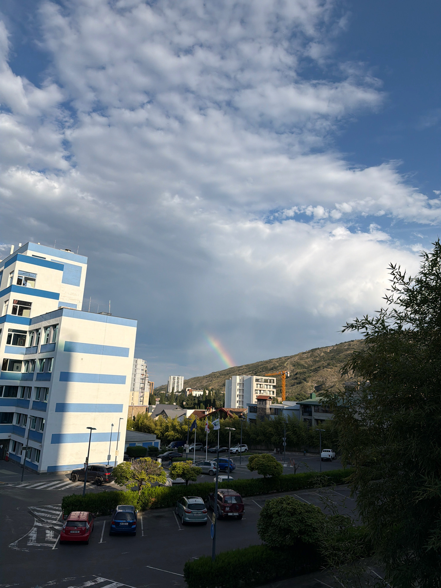 The image shows a residential area in Tbilisi, Georgia, with a rainbow visible in the sky. In the foreground, there are several parked cars and some greenery. A modern building with blue and white stripes is prominently featured on the left side of the image. In the background, there are more buildings, a hill, and a construction crane. The sky is partly cloudy with patches of blue sky. The lighting suggests it is either early morning or late afternoon.