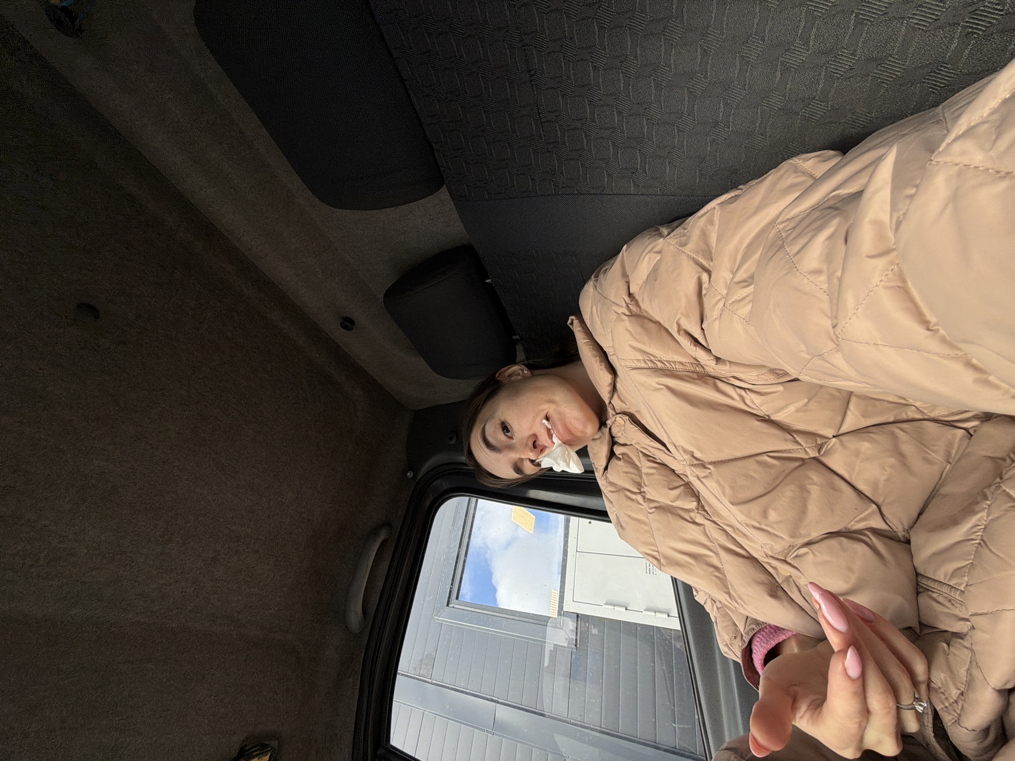 A young woman wearing a tan puffer jacket is seated in a vehicle, possibly a car or truck, with a tissue in her mouth. The interior of the vehicle is predominantly gray. Through the open window, a modern building with gray siding is visible. The sky is partly cloudy. The location is possibly in Izhevsk, Russia. The woman's hands are clasped in front of her, and she is wearing a ring.