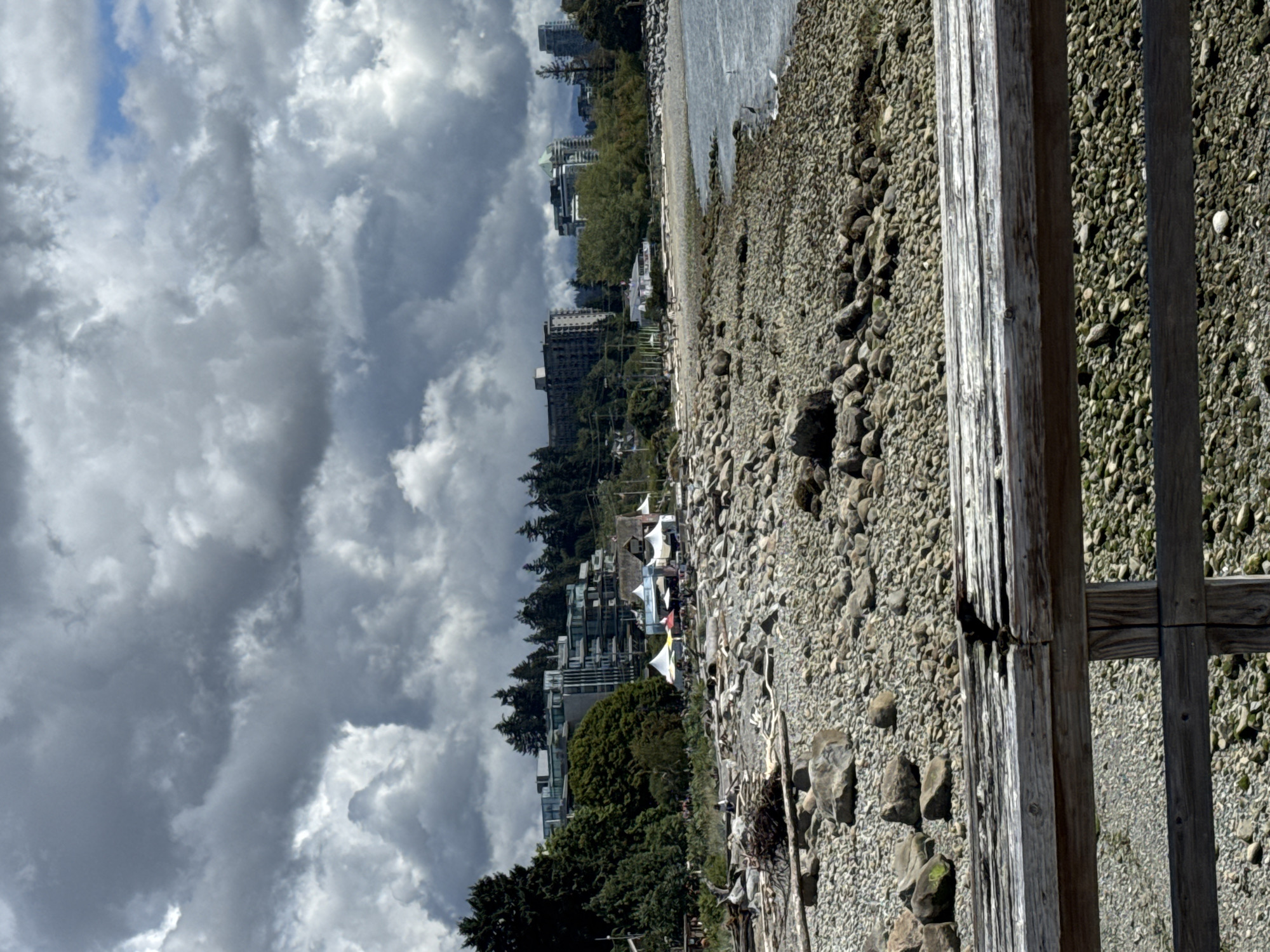 This is a medium-shot, eye-level photograph of a cloudy day at a beach in West Vancouver, Canada, likely Ambleside Beach, given the distinctive rocky shore and backdrop of apartment buildings. In the foreground, a weathered wooden railing runs horizontally across the bottom of the frame, with the texture of aged wood and weathered grain visible. Beyond the railing, a vast expanse of smooth, grey, and tan pebbles covers the beach. Scattered amongst the pebbles are larger rocks and driftwood, some of which are bleached white and others darker.
The middle ground features the shoreline, where the water meets the rocky beach. A few people are visible, appearing as small figures near some tents and structures set up on the beach. These structures include white tent-like canopies, some with colored accents, suggesting a temporary event or market. A building with a thatched roof and several other smaller buildings are also present. Further back, a line of lush green trees forms a dense forest, separating the beach area from a series of modern apartment buildings that rise into the frame. These buildings are multi-story structures with glass balconies and clean architectural lines. In the distance, behind the apartment buildings, faint outlines of mountains can be seen through the haze and clouds. The sky is dominated by dramatic, heavy grey clouds, with patches of brighter sky peeking through, indicating a dynamic and overcast weather condition. The sea is a muted grey-blue, reflecting the cloudy sky. The overall impression is a coastal urban scene under a moody sky.