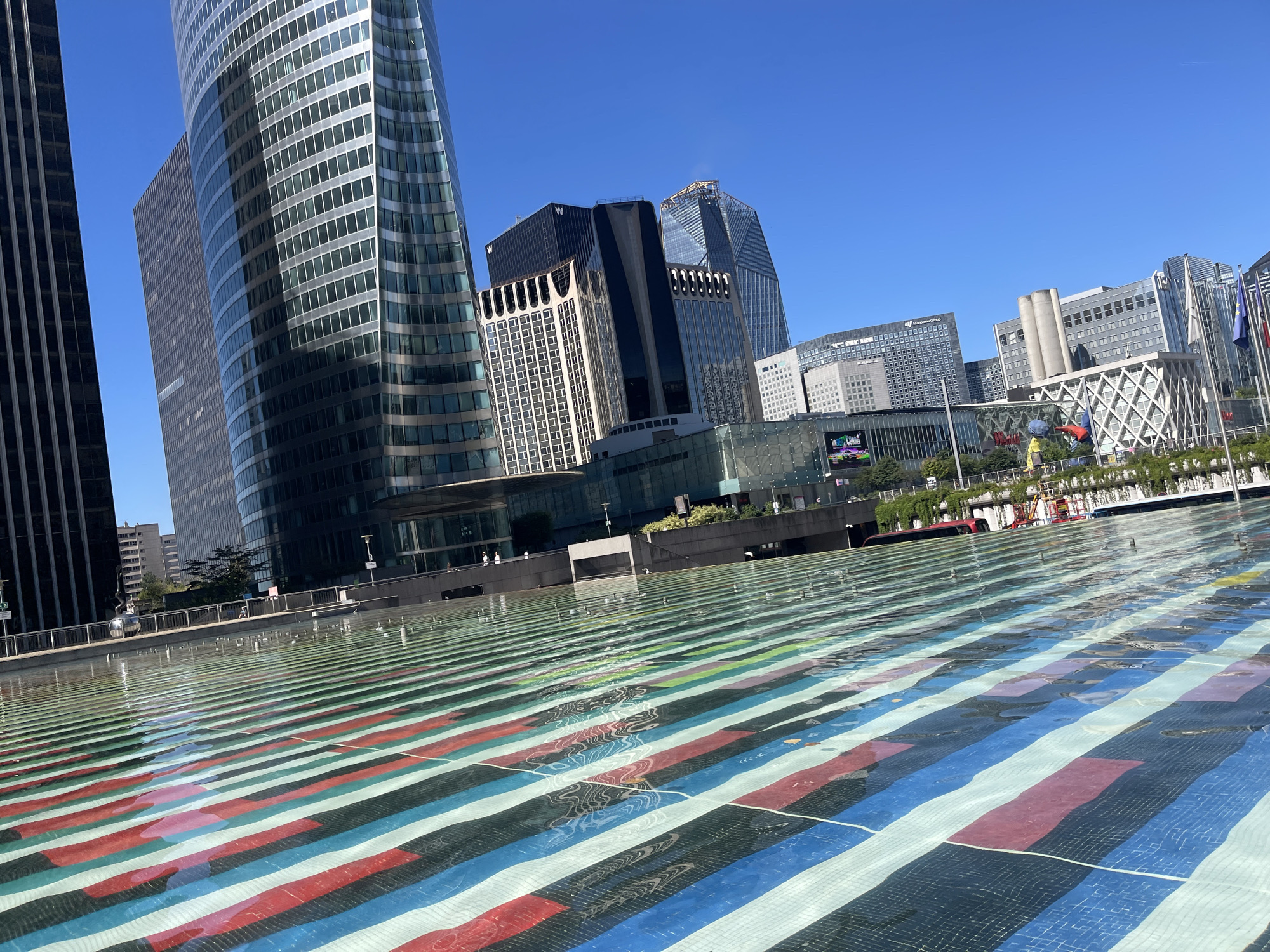 The image shows the outdoor fountain area in front of the skyscrapers of La Défense business district in Puteaux, France. The fountain's bottom is tiled with parallel stripes of red, blue, green, and white. The water refracts the sunlight, creating shimmering patterns across the stripes. Several modern skyscrapers with glass facades dominate the background, reflecting the clear blue sky. One of the skyscrapers on the left is a very tall, curved building with a grid pattern of windows. To its right is a dark-colored, rectangular skyscraper. In the middle ground, a cluster of buildings includes a dark, uniquely shaped skyscraper with a black diagonal band and a lighter-colored building with arched window details. Further back, a white, geometric-patterned building is visible, along with other office buildings. In the far background, several flags are displayed. The overall environment appears to be an urban plaza on a sunny day.
