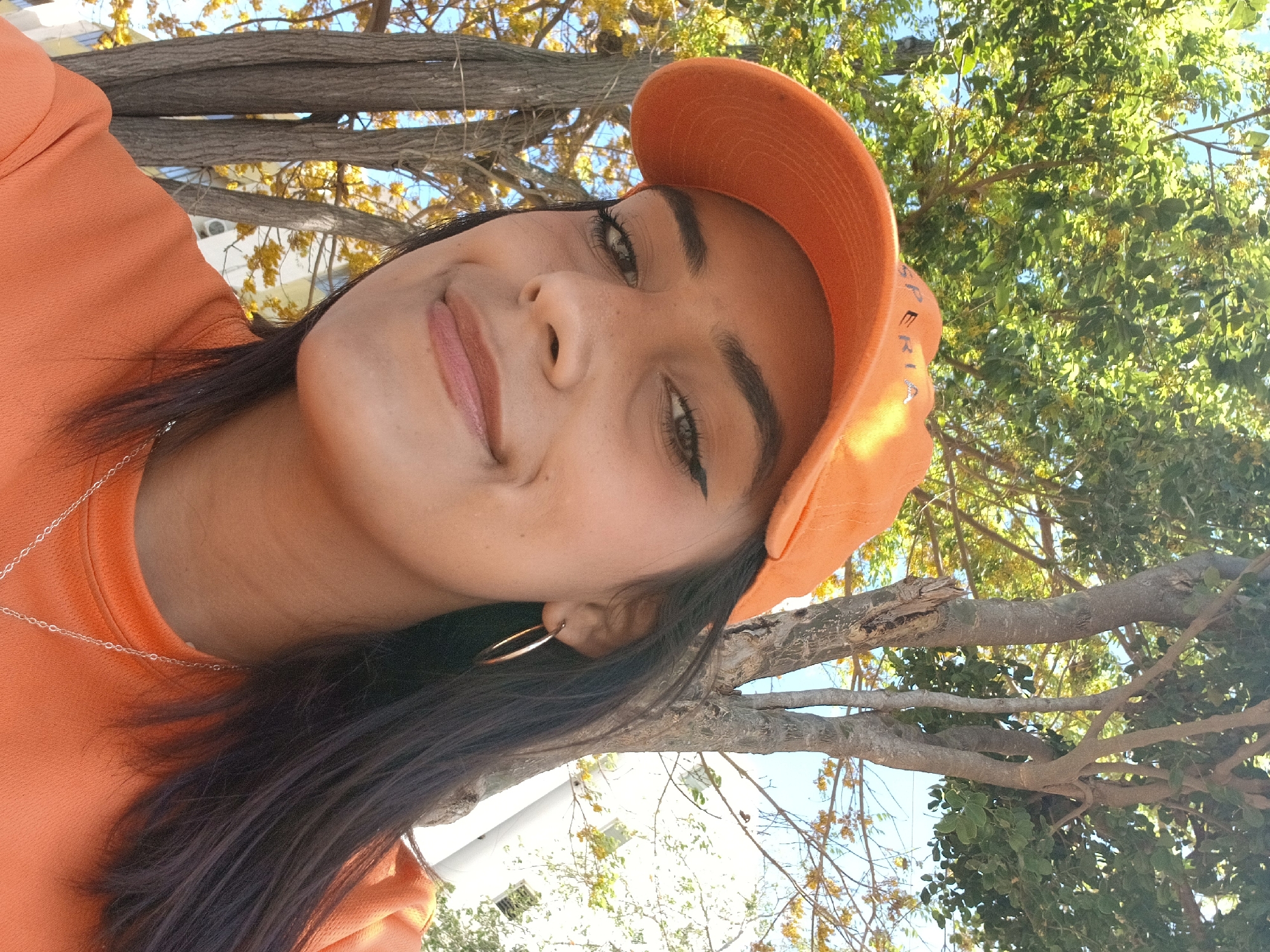 A woman wearing an orange top and a matching orange cap is taking a selfie outdoors. She is facing the camera at an angle, with her head tilted slightly. She is wearing a silver necklace and gold hoop earrings. The background consists of trees and foliage, and a building can be seen partially obscured by the tree branches. Based on the tags, this photo was possibly taken in Altagracia, Venezuela.