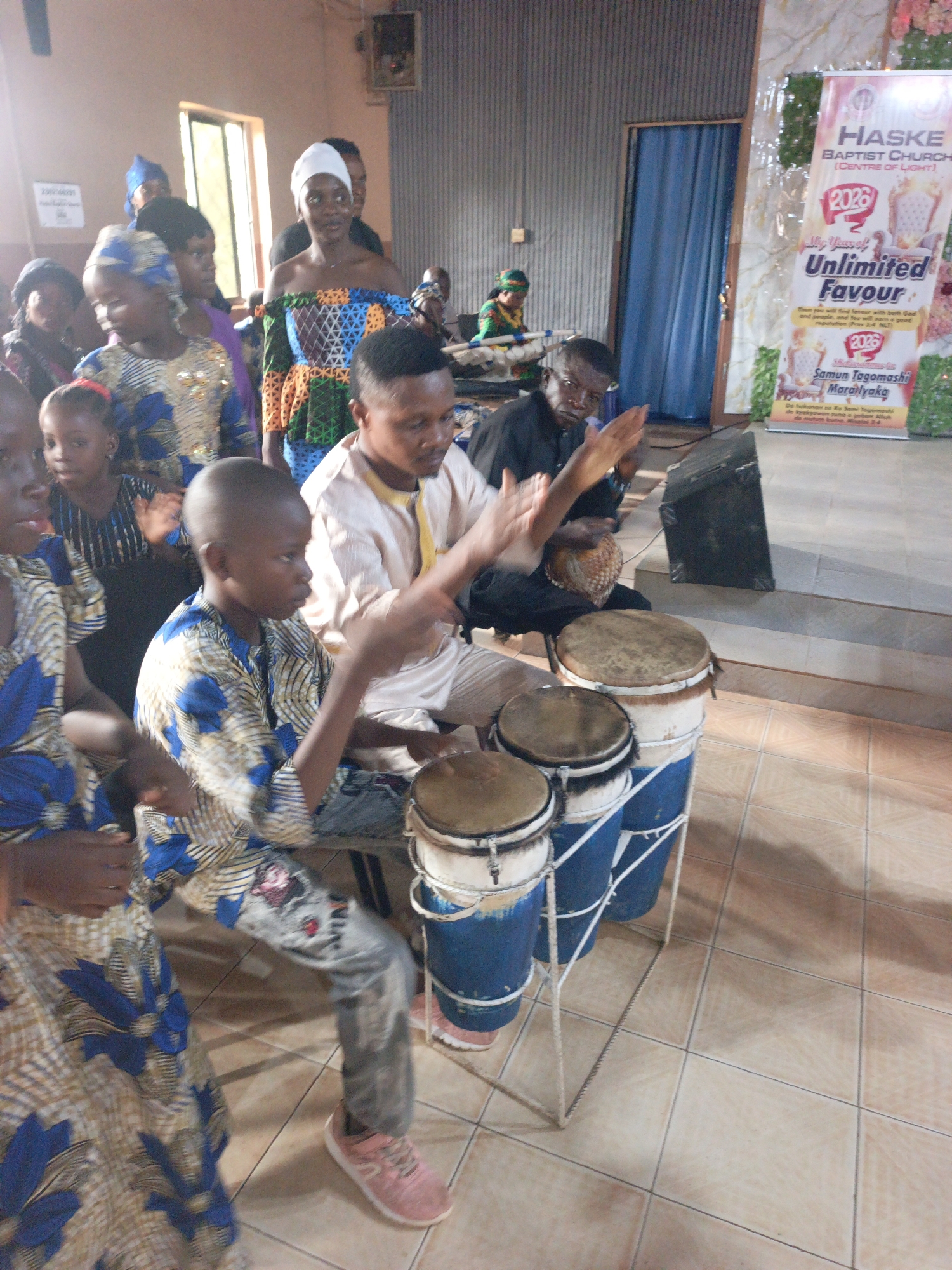 The photo captures a scene in Akwanga, Nigeria, likely a church or community hall. Two men and a young boy are seated, actively playing drums, while others stand around watching. The drums are a set of three stacked conga-style drums, painted blue, supported by a white metal frame. A man sits behind them playing a smaller drum. The drummer has a darker complexion and is wearing a black shirt. The boy on the left is wearing a patterned blue and gold top, grey jeans, and pink sneakers. Behind them, a group of people are standing and watching, some wearing traditional African clothing. A poster in the background reads "HASKE BAPTIST CHURCH" with the text "Unlimited Favour 2026" written below. The room has tile flooring, with a blue curtain hanging on the right side of the frame. A window is also visible in the background, letting in natural light.