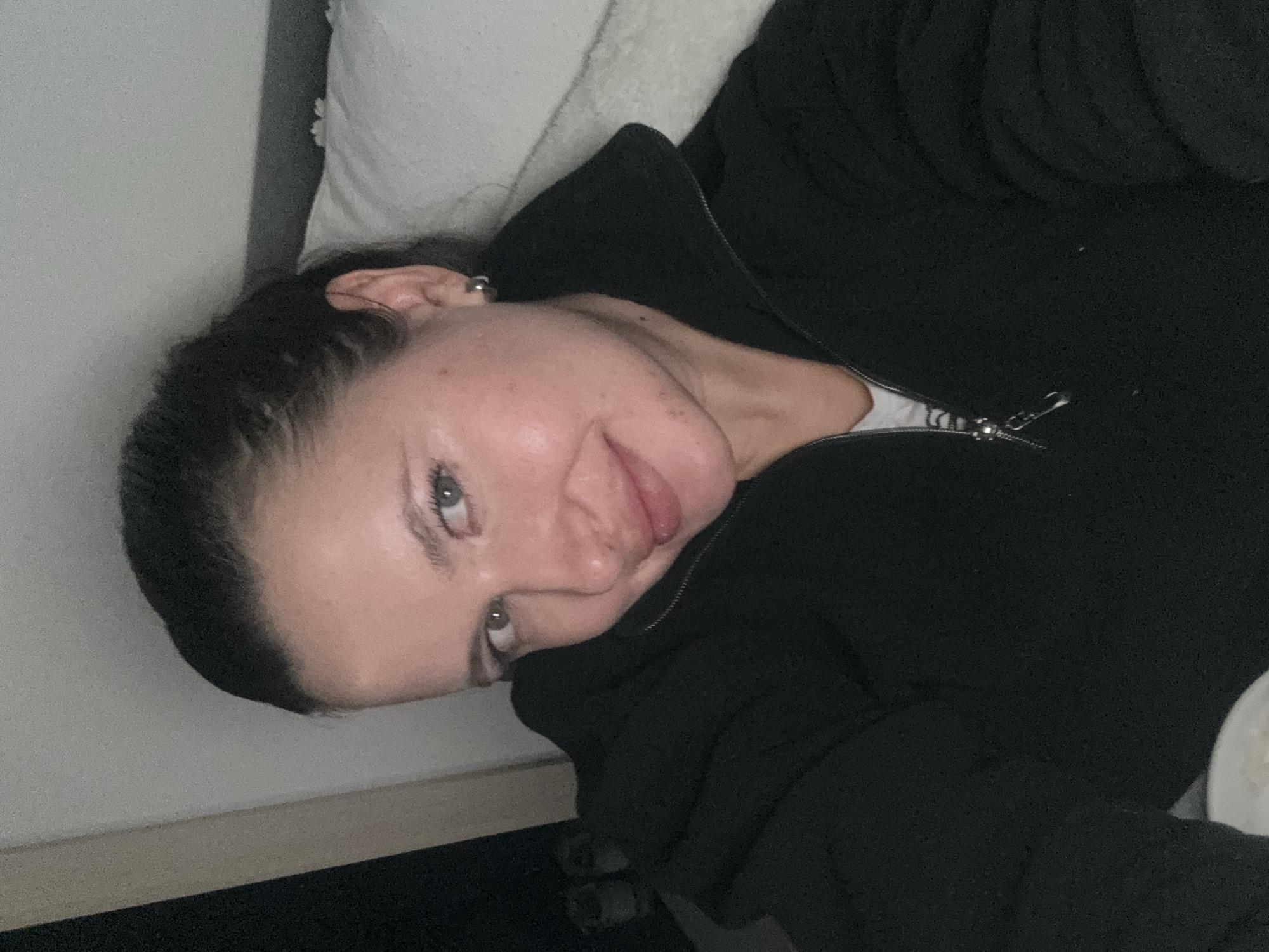 The media file displays a close-up portrait of a young woman, rotated 90 degrees clockwise, showing her lying down indoors. She is positioned on her side, resting her head against a white pillow. Her dark hair is pulled back, and she wears a small stud or hoop earring in her visible left ear. Her eyes are light-colored, and she offers a subtle, relaxed smile with her lips slightly parted, looking directly or slightly past the camera.
She is dressed in a dark, likely black, zipped-up jacket or hoodie, with a white garment visible underneath where the zipper is slightly open. The metallic zipper pull is clearly visible. The background consists of the white pillow behind her head and a dark surface, possibly a headboard or wall, to the upper left.
The lighting appears soft and diffused, characteristic of an indoor setting, possibly during the evening or under artificial light. The overall impression is one of casual comfort or a relaxed moment. There are no visible location cues within the image itself to confirm the provided location context of Akulovo (Gp Kubinka), Russia, nor is there any visible text.