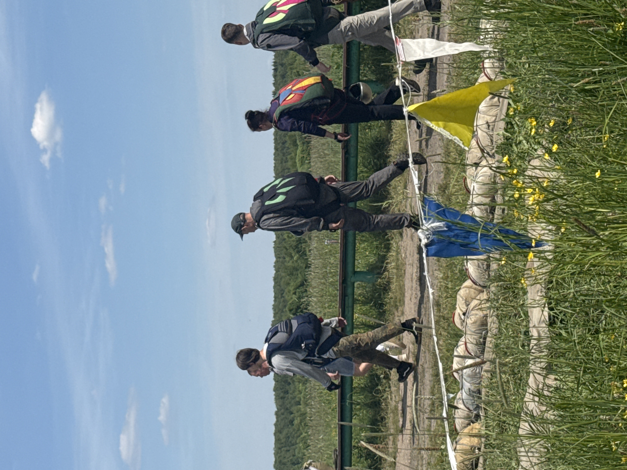 The image depicts an outdoor daytime scene at an unknown location. Under a clear blue sky with scattered white cumulus clouds, a group of five individuals is walking in a loose single file from left to right on a dirt path. All are equipped with large, specialized backpack-like rigs, consistent with skydiving parachutes.
From left to right:
1. A person is partially visible on the far left, wearing a light-colored top.
2. A male in a light-grey long-sleeved shirt, camouflage pants, and dark shoes, with a dark blue parachute rig strapped to his back.
3. A male wearing a dark cap, grey long-sleeved top, dark grey pants, and black shoes, carrying a black parachute rig with prominent bright green angular designs.
4. A female in a dark purple long-sleeved top, dark pants, and black shoes, carrying a multi-colored (red, yellow, blue, green) parachute rig, with a silver helmet visibly attached to the harness below the main pack.
5. A male in a grey jacket, light-colored pants, and dark shoes, carrying a dark green/black parachute rig with bright yellow/neon green angular designs.
Behind the individuals, a dark green metal barrier or railing extends horizontally across the midground. Beyond this barrier, a vast field of tall green crops, possibly wheat or similar, stretches towards a dense forest line on the horizon. The foreground is covered with tall green grass, interspersed with numerous small yellow wildflowers, likely buttercups. On the ground amidst the grass, several large, cylindrical objects wrapped in light-colored canvas, possibly rolled-up parachutes or equipment bags, are laid out. A white rope or cord is strung horizontally across the lower part of the foreground, from which a blue, a bright yellow, and a white rectangular flag are suspended. The scene suggests activities related to aviation or outdoor sports.