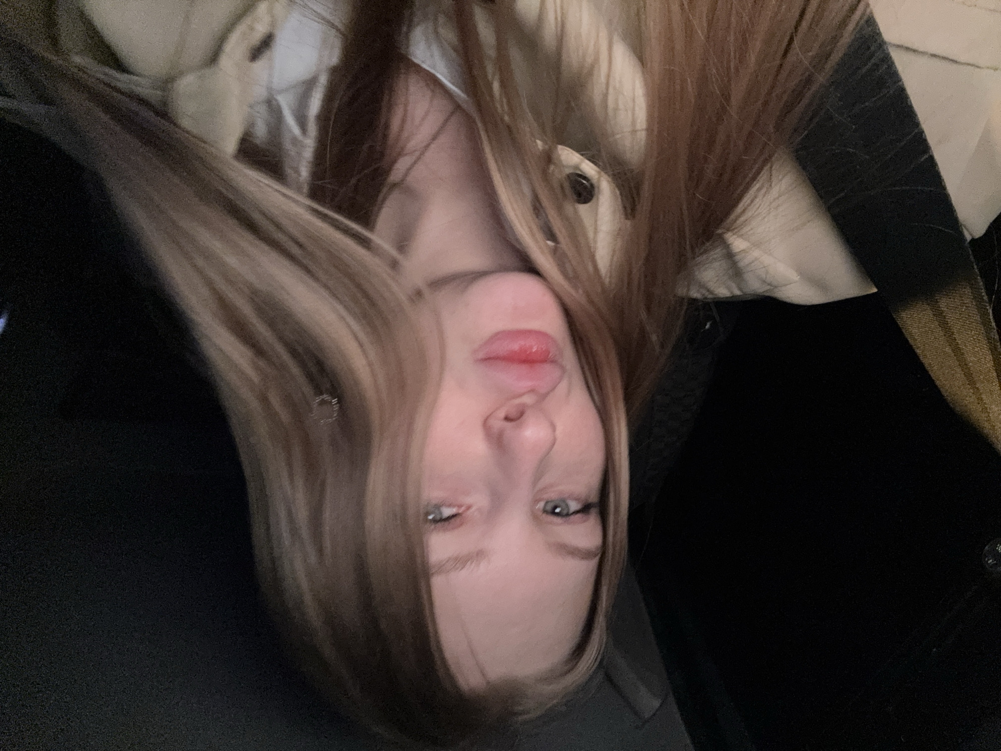 A young woman with light skin, blue eyes, and blonde-streaked brown hair is seen in an indoor setting, likely a vehicle due to the presence of a seatbelt strap. She is wearing a light-colored jacket or coat. The image is taken at an unusual angle, causing her face to appear upside down. The location in the city of Perm, Russia cannot be determined from the visual information provided.