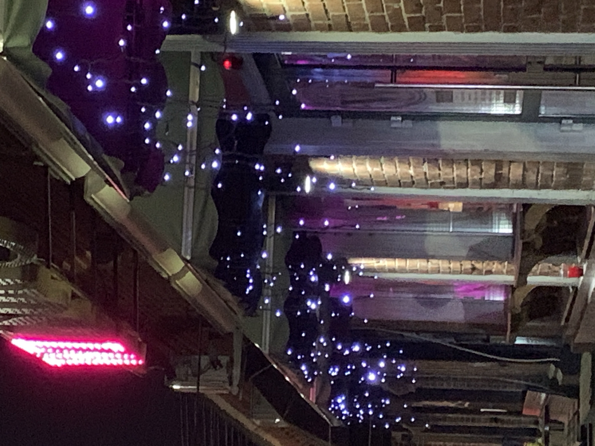 The image shows the exterior of a building at night, likely a business, decorated with string lights. A prominent, vertical pink neon light is visible on the upper left side. Numerous small, blue-white string lights are draped across the awnings and around the windows. The building appears to be constructed of brick. There are windows or doorways with reflective surfaces, and some are partially obscured by clear or opaque coverings. The scene is dimly lit, with the string lights providing the primary illumination. There are no people clearly visible in the image. The environment suggests an urban setting, possibly a cafe, bar, or shopfront. The exact location is unknown.