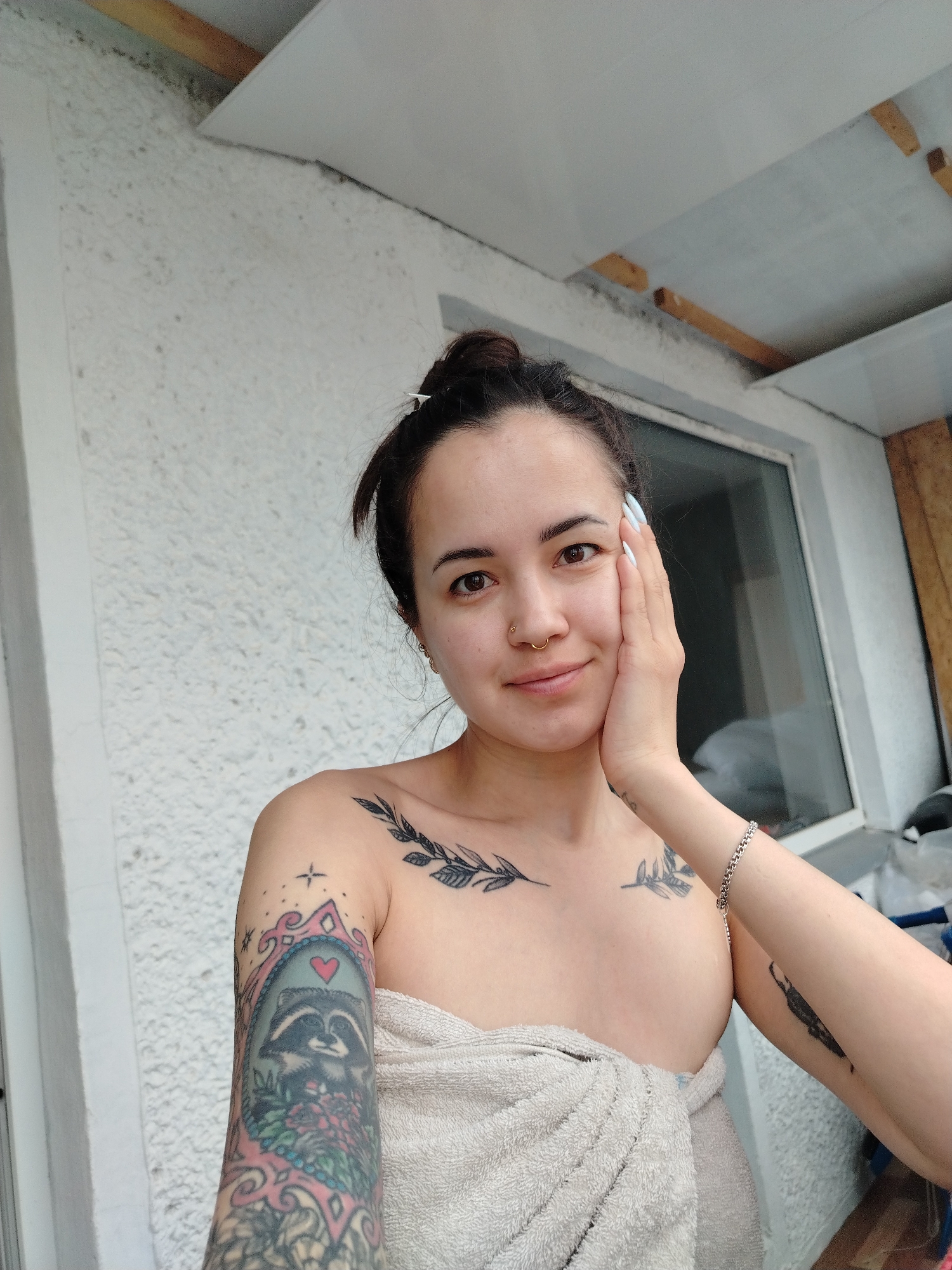 A young woman, presumably in Noviy, Russia, takes a selfie in what appears to be an outdoor or semi-outdoor setting, possibly a balcony or porch. She has dark hair tied up in a bun, brown eyes, and a subtle, content smile. A gold hoop nose ring adorns her septum. Her fingernails are painted a light color, and she wears a silver chain bracelet on her right wrist. She is wrapped in a light-colored towel around her torso, suggesting a post-shower or relaxed state, and gently touches her cheek with her right hand.
Her left upper arm and shoulder are prominently covered by a large, colorful tattoo depicting a raccoon holding flowers within an ornate, framed design. A black leafy branch tattoo is visible on her left collarbone, and another dark, abstract design or possibly a bird is tattooed on her right inner forearm.
The background features a textured white wall, likely stucco, and a large window reflecting natural, diffuse light, hinting at an overcast day. Above, a white ceiling or overhang is visible, with exposed wooden beams or supports, suggesting a functional or somewhat unfinished structure. The overall mood of the image is calm and personal. No text is visible.