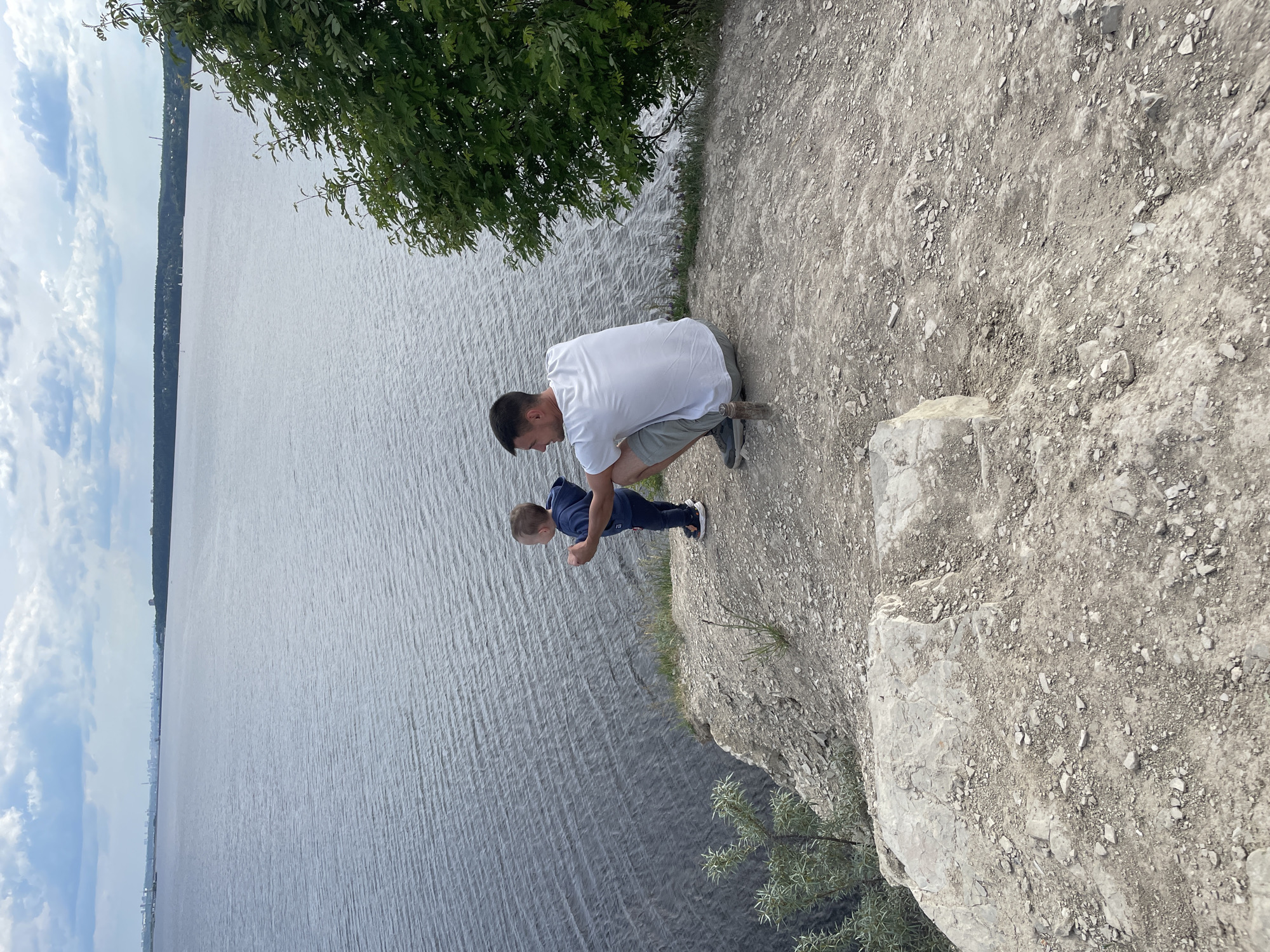 An eye-level shot captures a man and a young boy on a rugged cliff edge, looking out over a vast body of water under a partially cloudy sky. The man, wearing a white t-shirt and grey shorts, is crouched down on the rocky ground, his head turned slightly towards the boy. The boy, dressed in a blue long-sleeved top and dark trousers, stands next to him, also looking downwards, presumably at the water or what's below the cliff.
The cliff edge is composed of light grey, rough earth and scattered rocks and pebbles. A large green tree with leafy branches overhangs from the top left, partially framing the scene and casting some shade. Below the cliff, a wide expanse of water, likely a river or large lake, stretches across the frame, showing ripples on its surface. In the distance, on the far bank of the water, a thin strip of land is visible, featuring a line of trees and what appears to be a distant settlement or industrial structures. The sky above the distant horizon is bright with scattered white clouds.
The scene suggests a daytime setting, possibly late morning or early afternoon, with clear weather. The man and boy appear engaged in observing the view from their elevated position. There is no visible text in the image. The location is contextualized as Пятый километр, Russia, implying a scenic overlook.