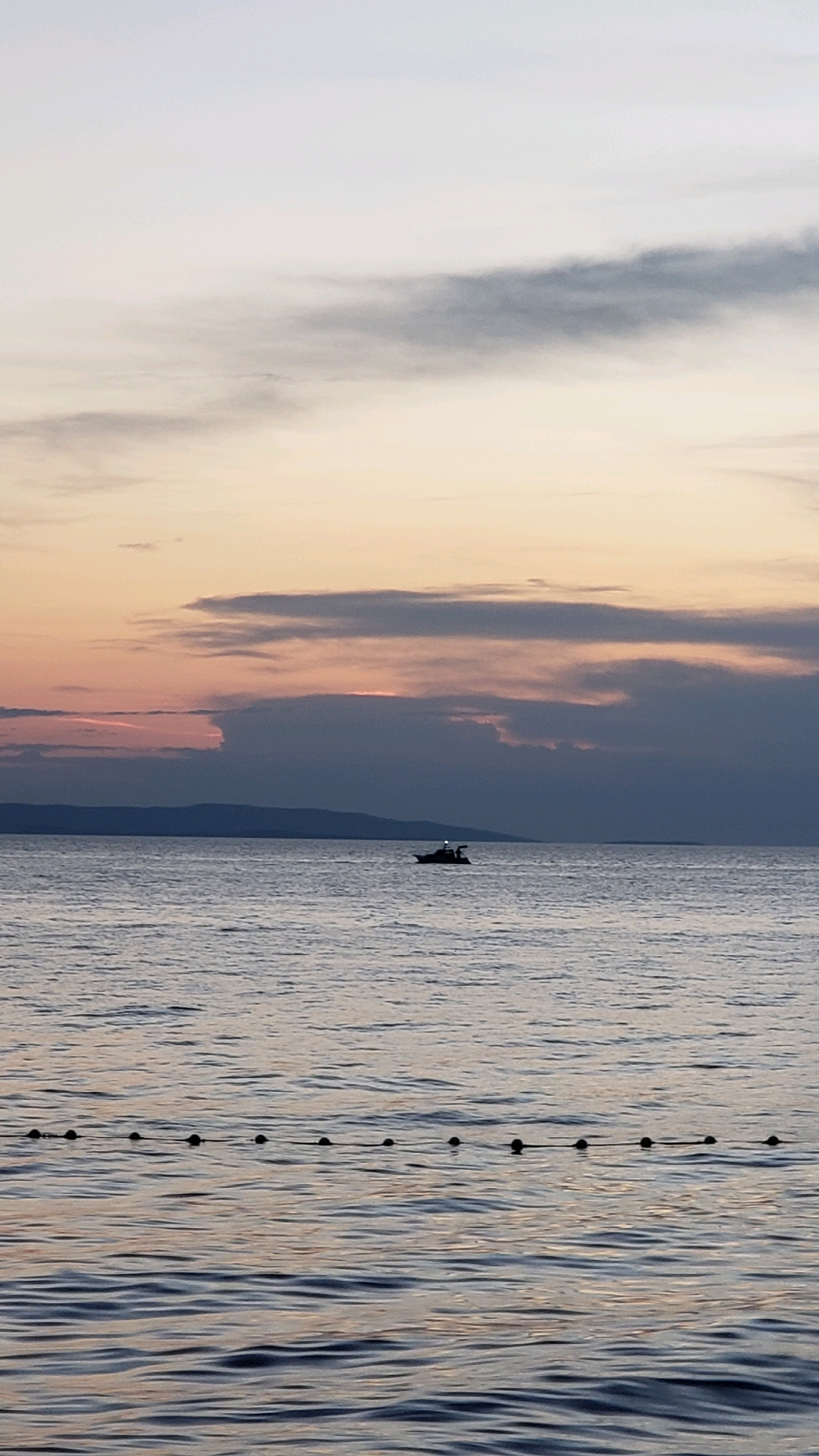 The image depicts a serene seascape at dusk in Općina Tučepi, Croatia. The sky is painted with soft hues of lavender, pink, and pale yellow, with wispy clouds stretching across the horizon. A dark, silhouetted mountain range forms the backdrop. In the middle ground, a small boat with a discernible cabin and a person or two on board navigates the calm, rippling water. A line of buoys, marked by dark spheres, floats on the water's surface in the foreground, suggesting a roped-off swimming area or a demarcation line. The overall atmosphere is tranquil and peaceful, capturing a moment of quiet solitude by the Adriatic Sea.