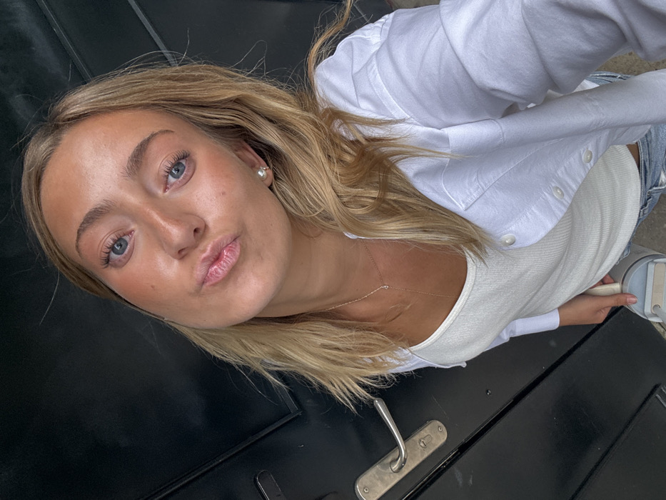 A female individual, approximately 20-30 years of age, with light brown skin tone, blue eyes, and long blonde hair, is depicted in a close-up, waist-up portrait. She is wearing a white ribbed tank top, an unbuttoned white collared shirt, and light-wash blue jeans. Her accessories include a delicate gold-colored lariat necklace with a small central clear stone and a single pearl stud earring in her right ear. Her lips are pursed, and she maintains direct eye contact with the camera. The subject is positioned in front of a dark, vertical surface, consistent with a painted or stained wooden door, identified by the presence of a silver-toned, horizontal door handle on the left side of the frame. The scene occurs in Mexico City, Mexico.