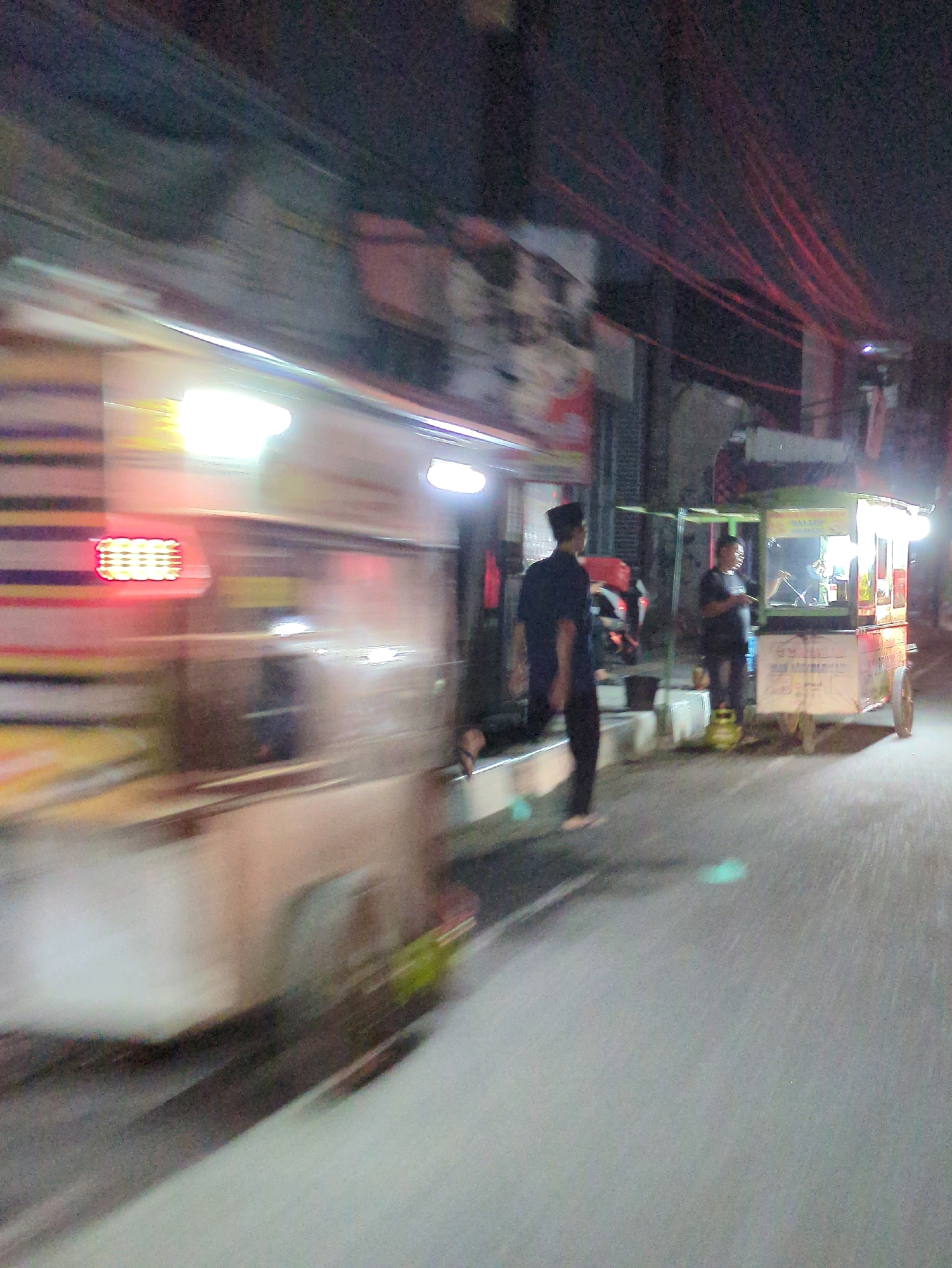 The image captures a nighttime street scene in Klodran, Indonesia, characterized by motion blur indicating movement. On the left, a vehicle or cart with horizontal stripes and bright lights is prominently visible. In the mid-ground, two individuals are present. One, wearing a dark cap and shirt, stands facing away from the camera. To the right, another individual is positioned near a food cart. The food cart, illuminated by lights, displays signage and appears to be operational. The environment suggests an urban setting at night, with overhead wires and buildings forming the backdrop. The ground surface is paved, and the overall lighting is low, with artificial light sources dominating the scene.