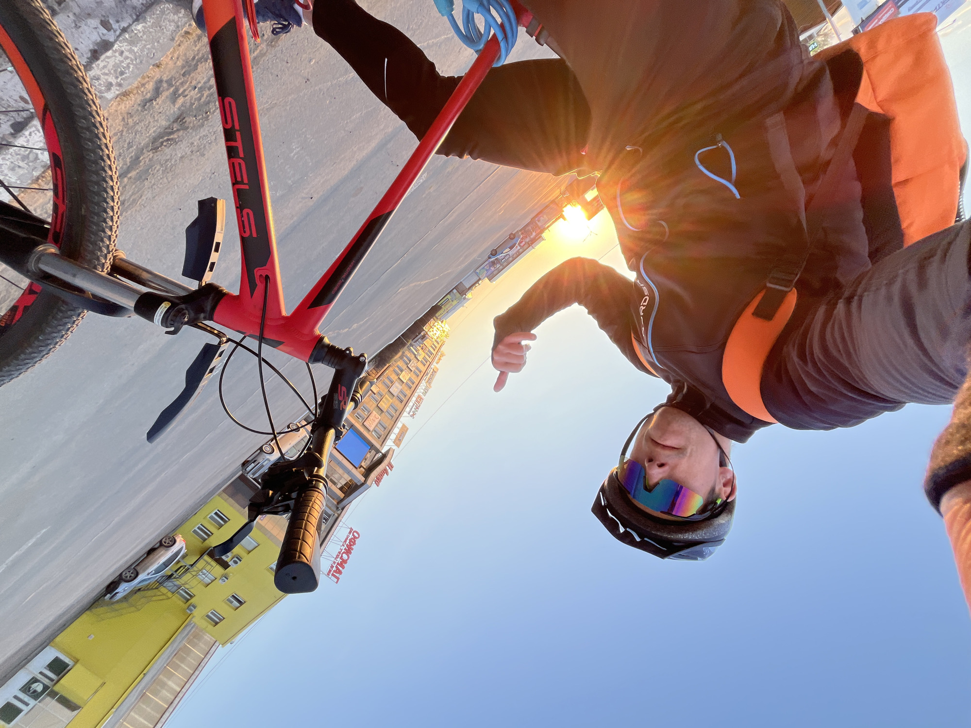 A cyclist is riding a Stels mountain bike on a road in Chaikovsky, Russia. The person is wearing a helmet and cycling glasses. The bike is red and black, and the background shows a yellow building with signage in Russian, possibly related to "OKHOMAT." The bright sun is also visible in the background. The person is pointing a finger downward toward the building and road, suggesting they are emphasizing a location.