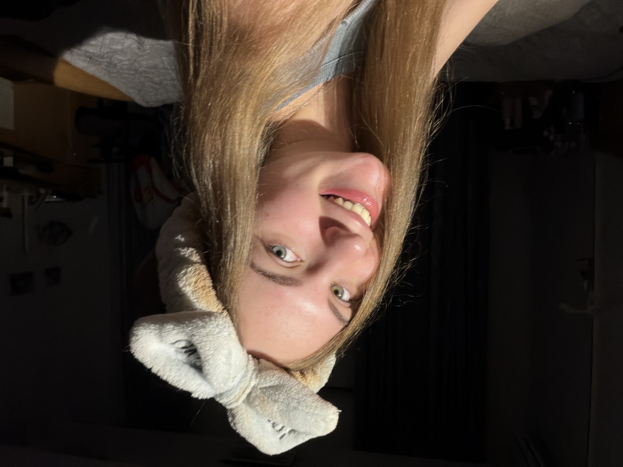 A young woman with long blonde hair and a fluffy headband with a bow smiles at the camera. She has striking blue-green eyes and her mouth is open in a wide smile, revealing her teeth and pink lips. She is wearing a light blue top and a silver necklace. The lighting suggests a warm, indoor environment, possibly a bedroom, with sunlight streaming in from the side, casting a shadow on the background. The background is dark, with hints of furniture and a bed. The image is a close-up portrait, focusing on the woman's face. The location is not specified in the image or the provided crops.