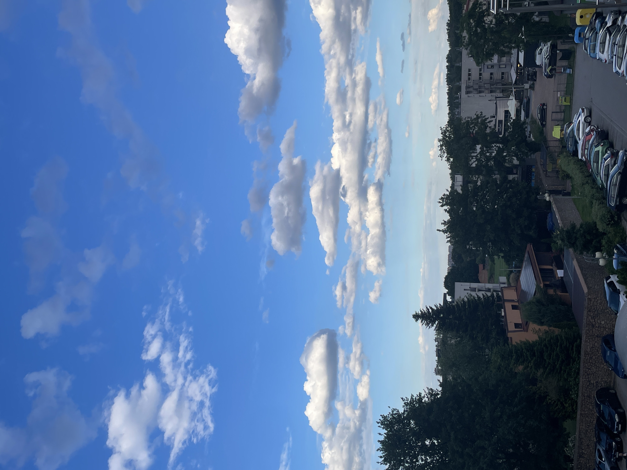The image is a high-angle shot of a residential area with a clear blue sky filled with white clouds. In the foreground, there is a parking lot with several cars. Behind the parking lot, there are trees, buildings, and more parked cars. The buildings appear to be apartment complexes and houses. The overall scene suggests a suburban or urban environment during the daytime.