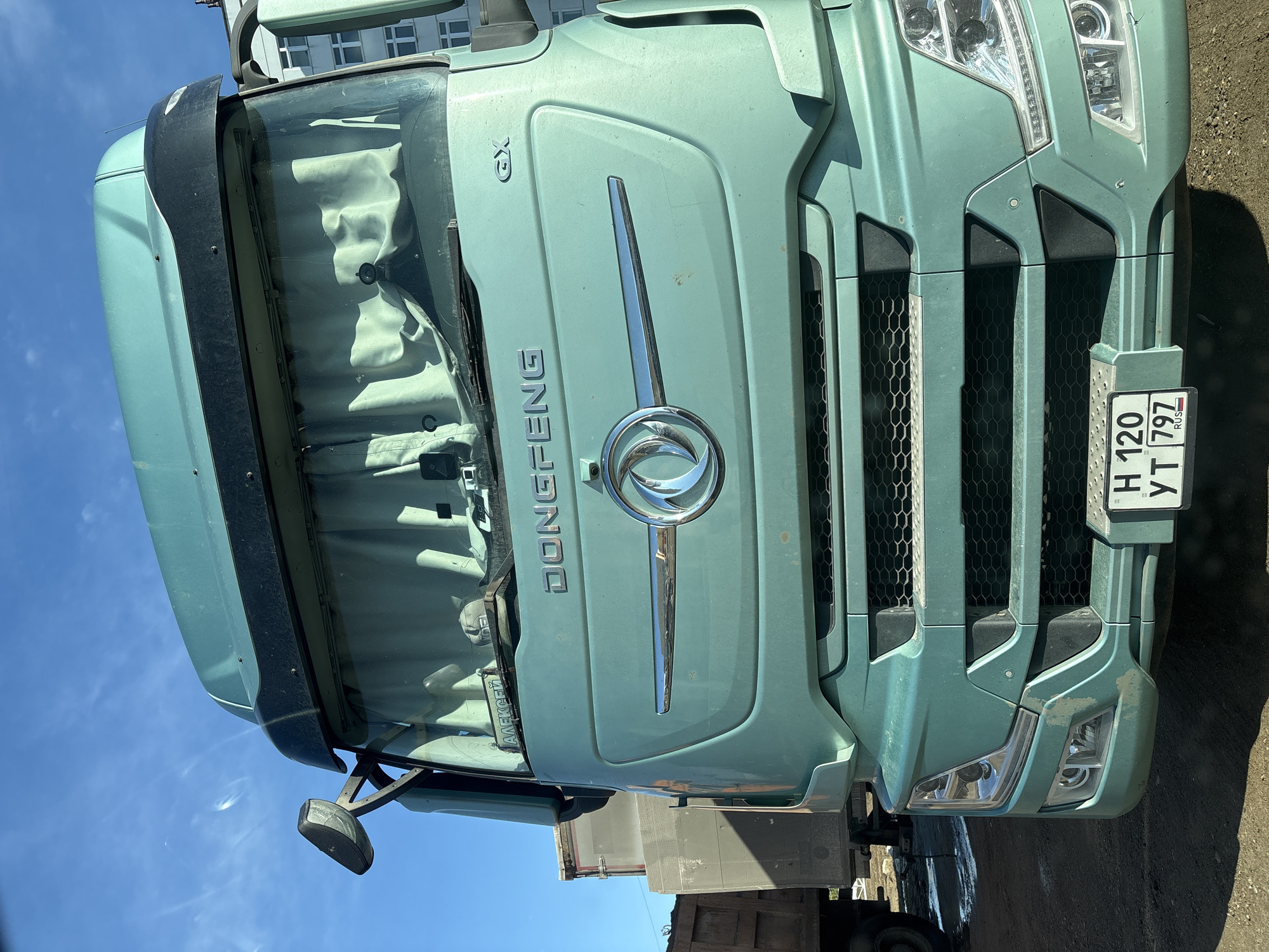 The image shows the front of a green Dongfeng truck. The license plate reads "H 120 YT 797 RUS." The location is Chaikovsky, Russia, based on the license plate. The truck has a silver logo and grill. The windshield has curtains inside. The truck appears to be parked on a dirt or paved surface. The sky is visible in the background.