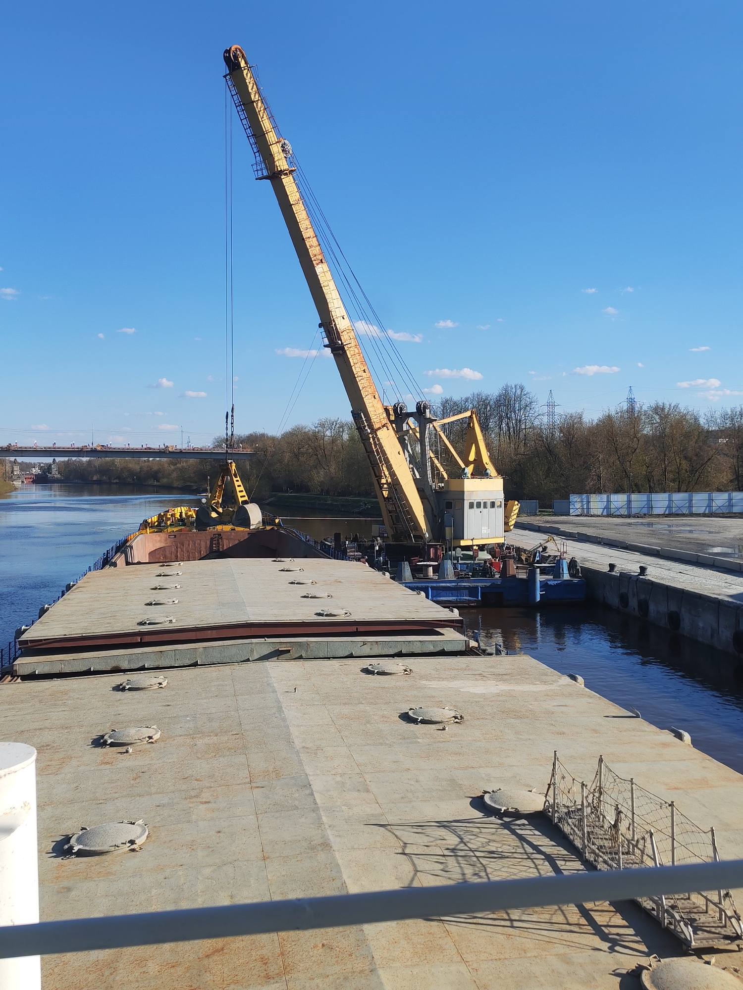 A large, flat-deck barge is docked alongside a concrete quay, with a large, yellow crane dominating the scene. The crane is positioned on the barge's stern. The crane's boom is angled upwards, with its hook and cables visible. Further up the barge, is another smaller yellow crane in the foreground. The barge's deck is covered in large, circular hatches. A small bridge, partially obscured by trees and landscape, crosses a canal in the background. There are trees and industrial buildings along the banks of the canal. Overhead power lines are visible. The sky is blue with scattered white clouds. The waterway is the Yakhroma Canal, located in the city of Yakhroma, Russia. The scene appears to be an industrial area along the canal.