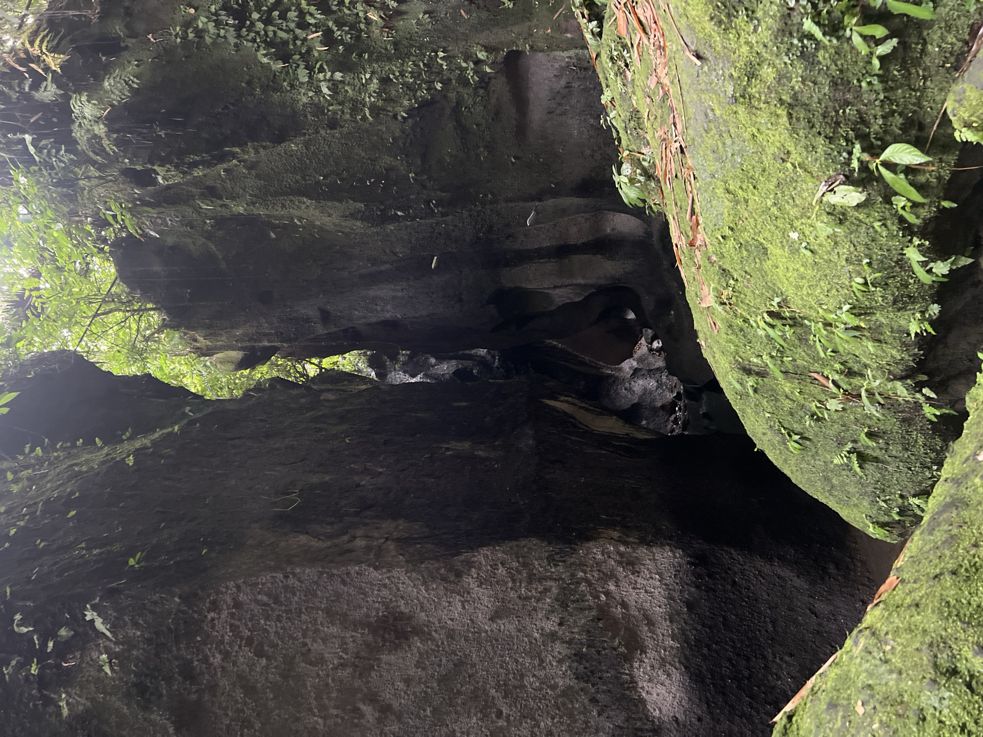 The image shows a narrow, dark canyon with rocky walls in Tembuku, Indonesia. The walls are covered in moss and vegetation. Light filters in from above, illuminating a small stream or body of water at the bottom of the canyon. There are no people visible.