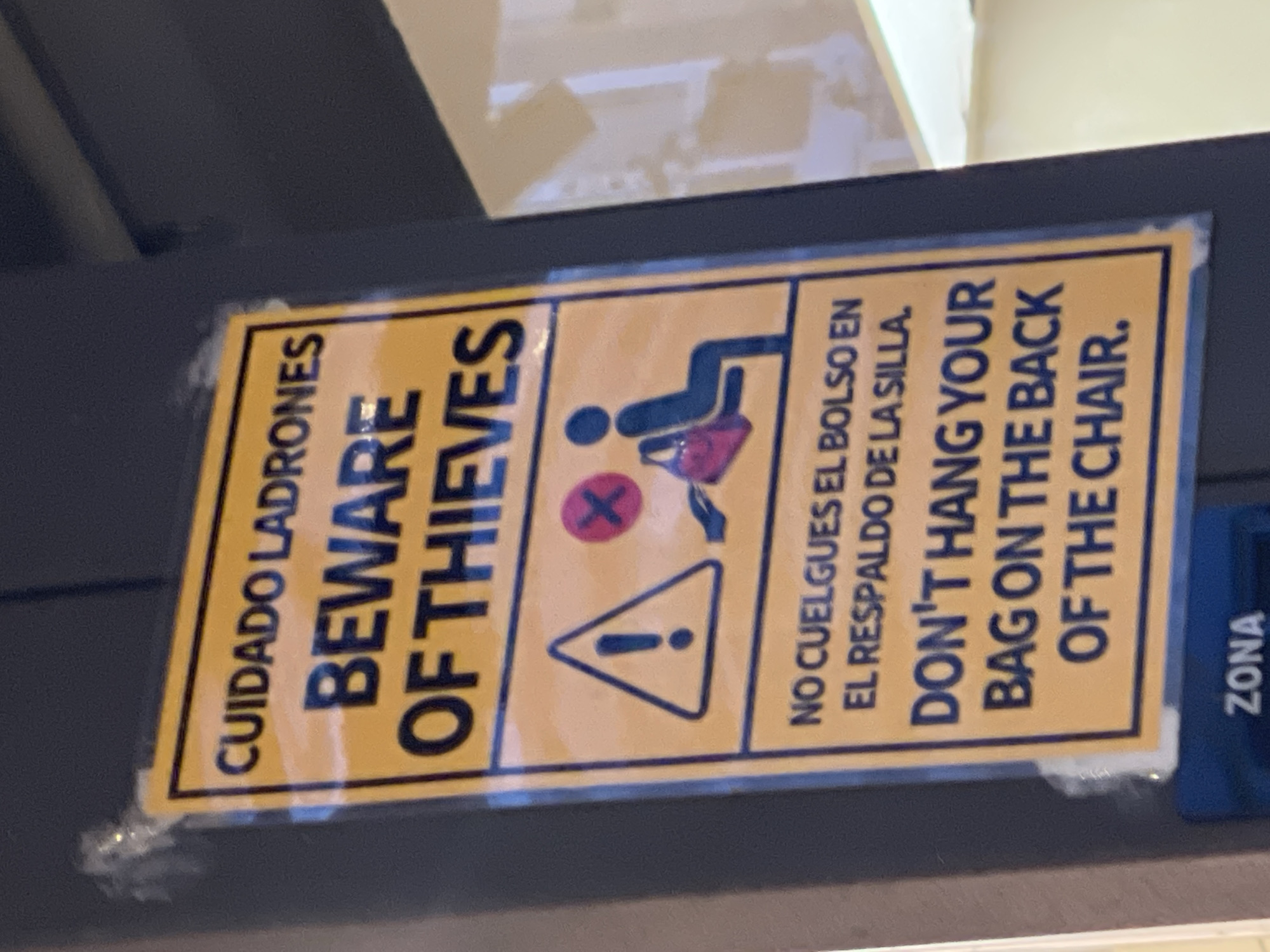A yellow sign with black lettering is affixed to a dark gray pole. The sign has a blue border and contains text in both Spanish and English, along with a warning symbol. The Spanish text at the top reads "CUIDADO LADRONES" and below that, "BEWARE OF THIEVES". Further down, it says "NO CUELGUES EL BOLSO EN EL RESPALDO DE LA SILLA" and "DON'T HANG YOUR BAG ON THE BACK OF THE CHAIR." To the left of the English text, a triangular warning symbol with an exclamation mark is depicted. To the right of this, a red circle with a diagonal line through it, superimposed over a pink bag, is shown next to a silhouette of a person sitting on a chair. The sign is slightly crumpled in the top left corner, where it appears to be attached with clear tape. Below the main sign, a dark blue sign with the word "ZONA" is partially visible. The background suggests an indoor environment, possibly a cafe or public waiting area, with blurred reflections of light and possibly furniture in the upper right. The presence of "ZONA" suggests this could be a designated area within a larger establishment, and given the multilingual warning, the location is likely a tourist-frequented area. Based on the Spanish text and the commonality of such warnings in tourist locations, it is plausible this sign is in a public space in Barcelona, Spain.