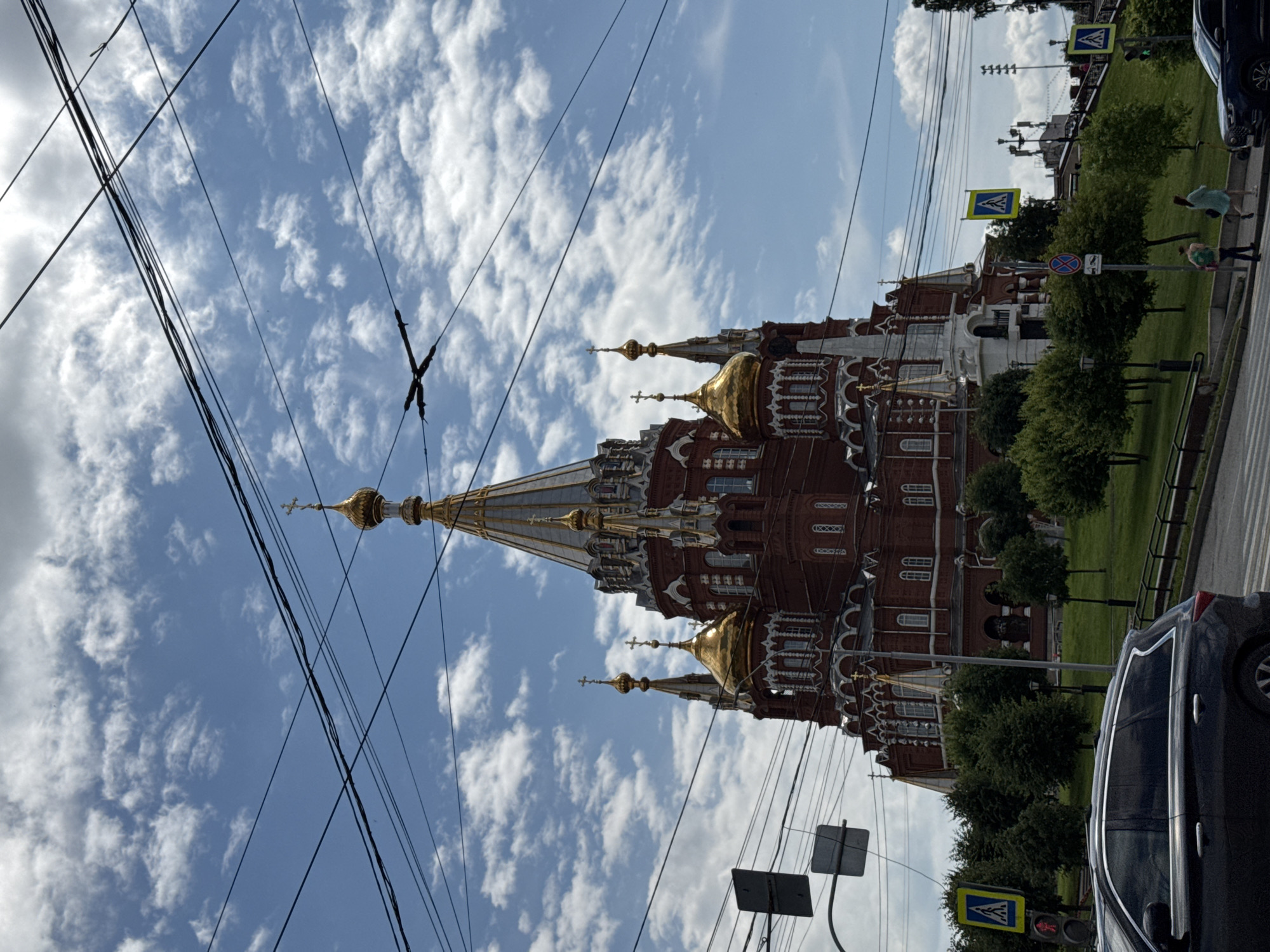 The image depicts the Alexander Nevsky Cathedral in Izhevsk, Russia, under a partly cloudy sky. The prominent red brick cathedral features multiple golden domes, with the central dome being the largest. The foreground shows a dark-colored SUV driving on a street with a pedestrian crossing. Traffic lights and street signs are visible. Green grass, trees, and a pathway are situated between the road and the cathedral, with several people walking along the pathway. Overhead, a complex network of power lines crisscrosses the sky, obscuring parts of the cathedral's spires and the clouds. The overall scene captures a typical urban environment with significant architectural elements and daily activities.
