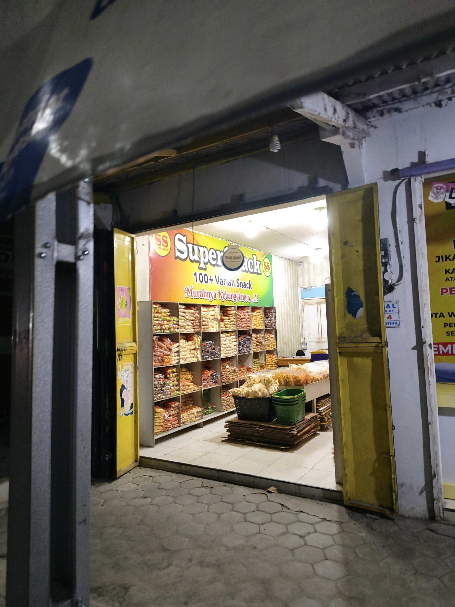 The image shows the entrance of a snack shop in Derekan, Indonesia. The shop is brightly lit and filled with shelves stocked with various packaged snacks. A large sign above the shelves reads "Super Snack 100+ Varian Snack Murahnya Kebangetan!!!". The entrance is framed by yellow doors, one of which is ajar. Outside the shop, the ground is paved with hexagonal tiles. In the foreground, there are grey metal pillars and a portion of the building's facade. The scene appears to be at night, judging by the artificial lighting.