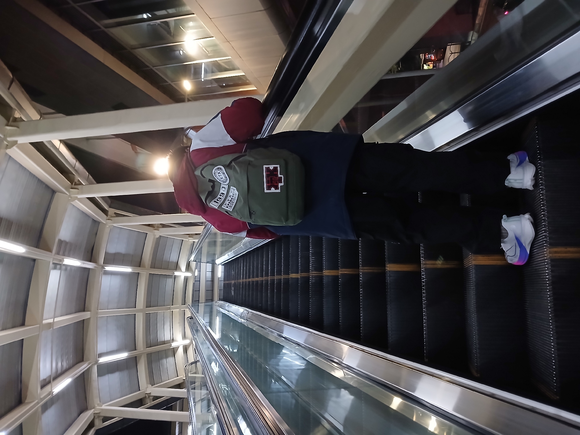 A person wearing a red and white jacket, dark blue pants, and white sneakers with blue and pink accents is riding an escalator. The person is also wearing a green backpack with patches. The escalator is in the city of Chongqing High-tech Zone, China, based on the provided context. The escalator has a glass barrier on both sides and is descending. The architecture surrounding the escalator includes white beams and a translucent ceiling. The escalator treads are black with thin yellow safety lines.