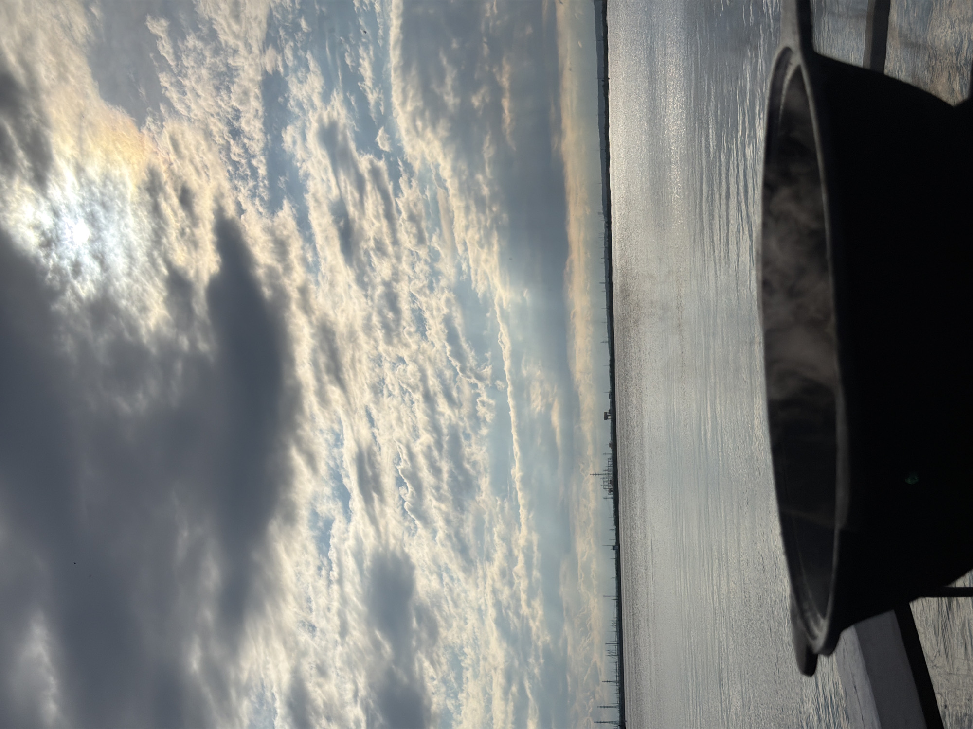 A wide shot captures a serene, overcast day by the water, with a dark, wide pot in the foreground, emitting a visible steam. The pot is positioned on a dark railing, suggesting it's on a balcony or deck overlooking a body of water. The water is calm and reflects the sky, which is filled with dramatic, layered clouds. Sunlight breaks through in some areas, creating shafts of light and a faint rainbow-like effect near the sun. In the distance, a faint industrial skyline is visible along the horizon, with numerous structures and towers, likely indicative of a port or industrial area. This scene appears to be located in Chaikovsky, Russia, a city known for its industrial landscape and proximity to the Kama River.