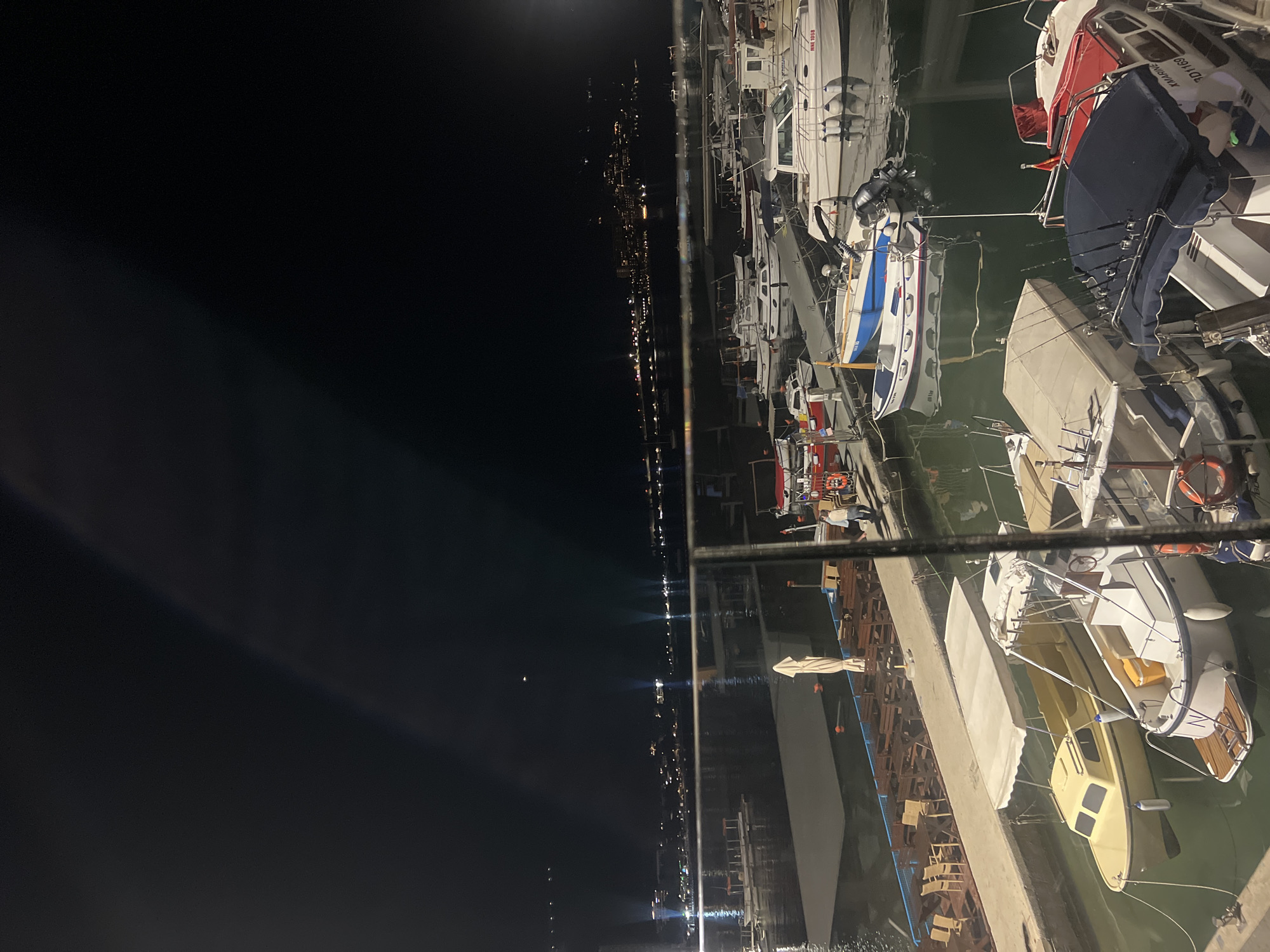 The image depicts a nighttime scene at a marina, likely in Budva, Montenegro, given the city lights visible across the water. Several boats are docked along a concrete pier. The boats are of various sizes and colors, including white, blue, red, and yellow. They are moored close together. The water appears dark, reflecting the city lights. The city lights create a bright horizon line. There is very little apparent activity. The scene suggests a calm evening at a port.