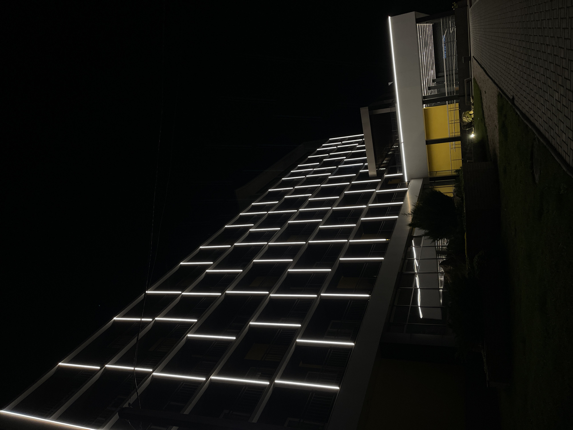 The image depicts the facade of a modern building at night, illuminated with linear lights arranged in a grid pattern. The building appears to have multiple floors, with some sections featuring yellow-colored walls visible through windows. A section of paved ground and a grassy area are adjacent to the building. The location appears to be Tiraspol, Moldova.