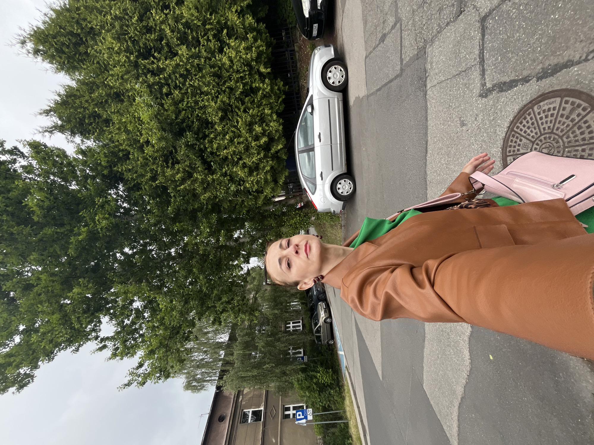 In an outdoor urban setting, a woman is captured in a selfie as she stands on a paved street, facing the camera. She is wearing a brown leather jacket over a green top and is holding a light pink shoulder bag. The background features a building with windows and several parked cars, including a silver sedan. Lush green trees and bushes line the street, and the sky is overcast. The woman's expression is neutral, and her hair is pulled back. She is positioned in the foreground of the image, with the urban environment extending behind her.