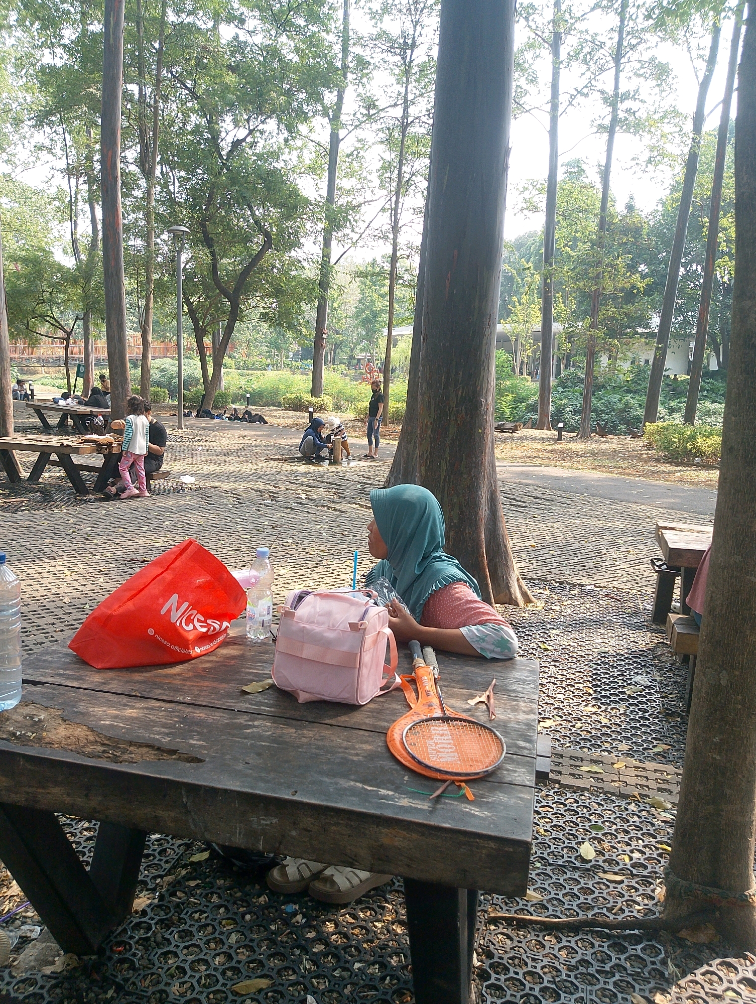 The image depicts a park scene in South Jakarta, Indonesia, on a sunny day. In the foreground, a wooden picnic table is laden with items. A large red bag with "Niceso" branding sits prominently, alongside a plastic water bottle and a pink backpack. An orange badminton racket lies on the table, suggesting recreational activity. A person wearing a turquoise hijab and a patterned shirt is seated at the table, looking to the right. The table is positioned on a textured grey ground surface surrounded by trees and other picnic tables in the background. In the middle ground, several people are visible, some seated at tables and others walking. The park appears well-maintained with lush green trees and foliage.