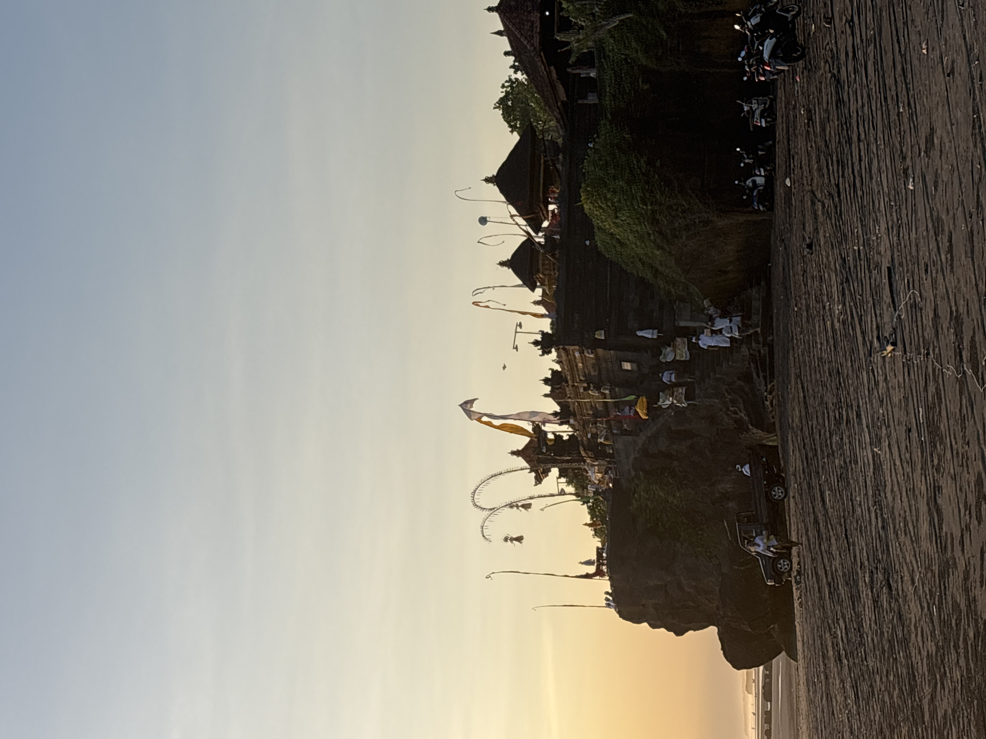 Captured in Kerambitan, Indonesia, the image presents a detailed scene at dusk. A traditional Balinese temple complex occupies a rocky promontory jutting into the sea. The temple structures are adorned with flags and ceremonial decorations. People in traditional white attire are gathered within the temple grounds. Motorbikes are parked atop the cliff, beside a wooden walkway. A Jeep, with occupants visible, sits on the cliff's edge, below the temple. The sky transitions from blue to sunset orange.