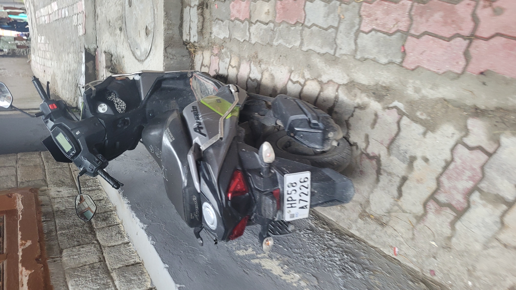 A black Suzuki Avenis scooter is parked on a wet, patterned concrete walkway next to a gray stone wall. The scooter's license plate reads "HP58 A7226". In the background, out of focus, there are people standing on a street. The scene appears to be in Vashisht, India.