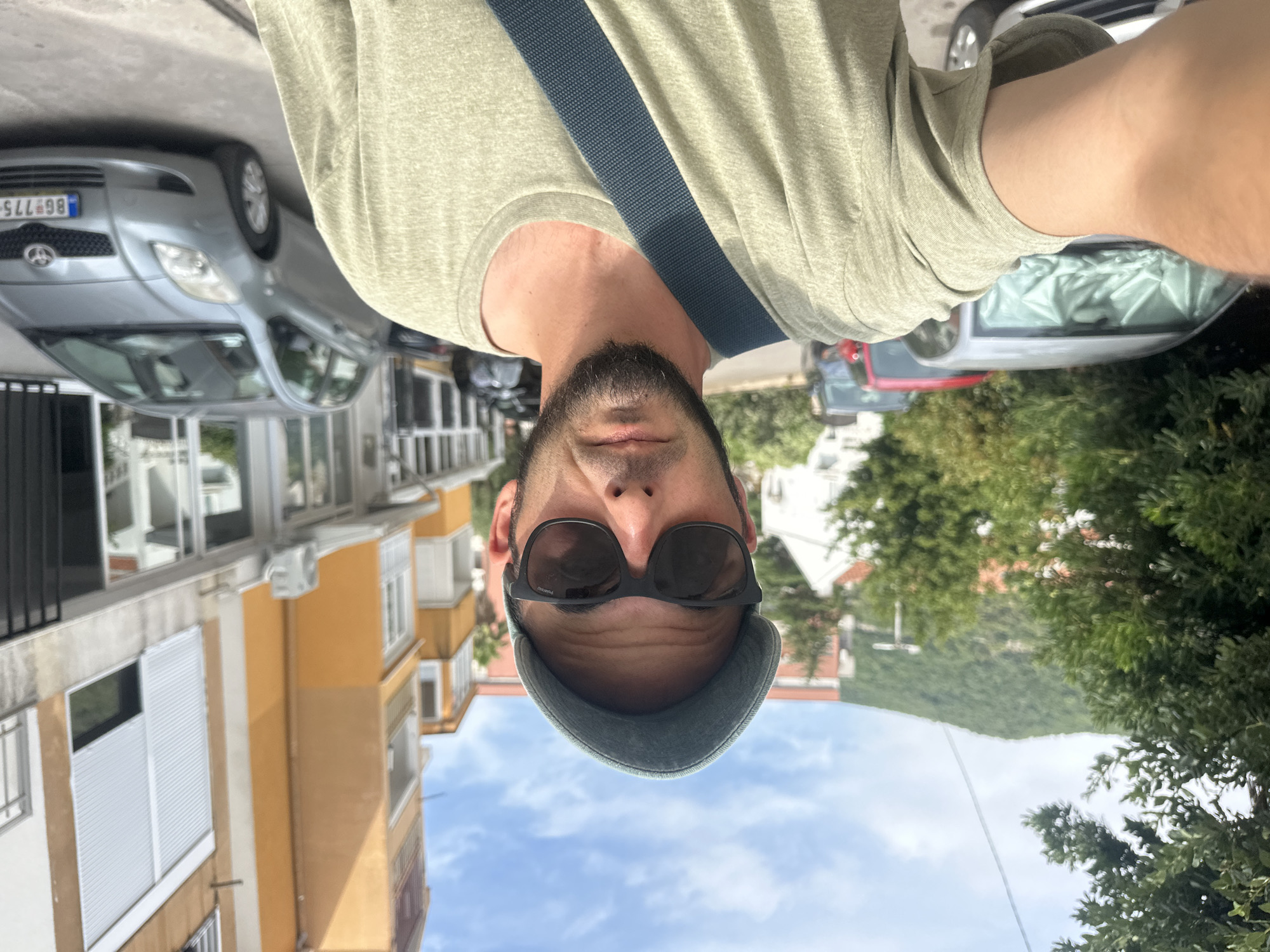 A man wearing sunglasses and a baseball cap takes a selfie on a street in Budva, Montenegro. He is wearing a light green t-shirt and a blue strap is visible across his chest. Behind him, several cars are parked on the street, and apartment buildings line the background. The sky is blue with some clouds, and green trees are visible.