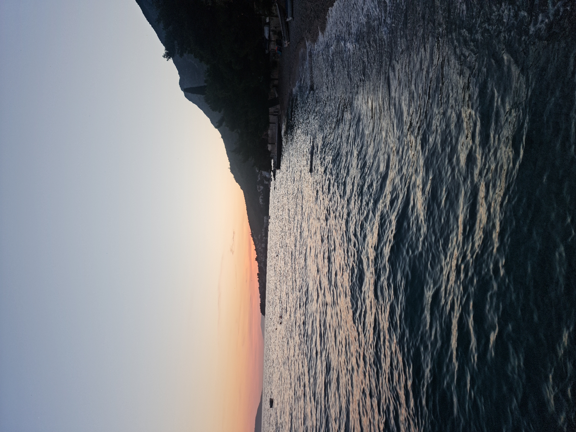 A vertical, low-angle shot captures the Adriatic Sea in Brist, Croatia, during dusk. The sky transitions from a pale blue at the top to a soft orange and pink near the horizon. The sea occupies the majority of the frame, with gentle waves lapping at a pebble beach.
On the beach, dark silhouettes of trees and vegetation line the base of a mountain that rises steeply on the right side of the image. The mountain is covered in dark foliage and has a rugged, natural appearance. A few scattered objects, possibly boats or debris, are visible along the shoreline. In the distance, a cluster of buildings suggests the presence of the village of Brist. The water is a deep blue-green, reflecting the dimming light of the sky. A solitary boat is anchored in the calm water on the left side of the frame. The overall atmosphere is serene and tranquil, indicative of a peaceful evening by the sea.
