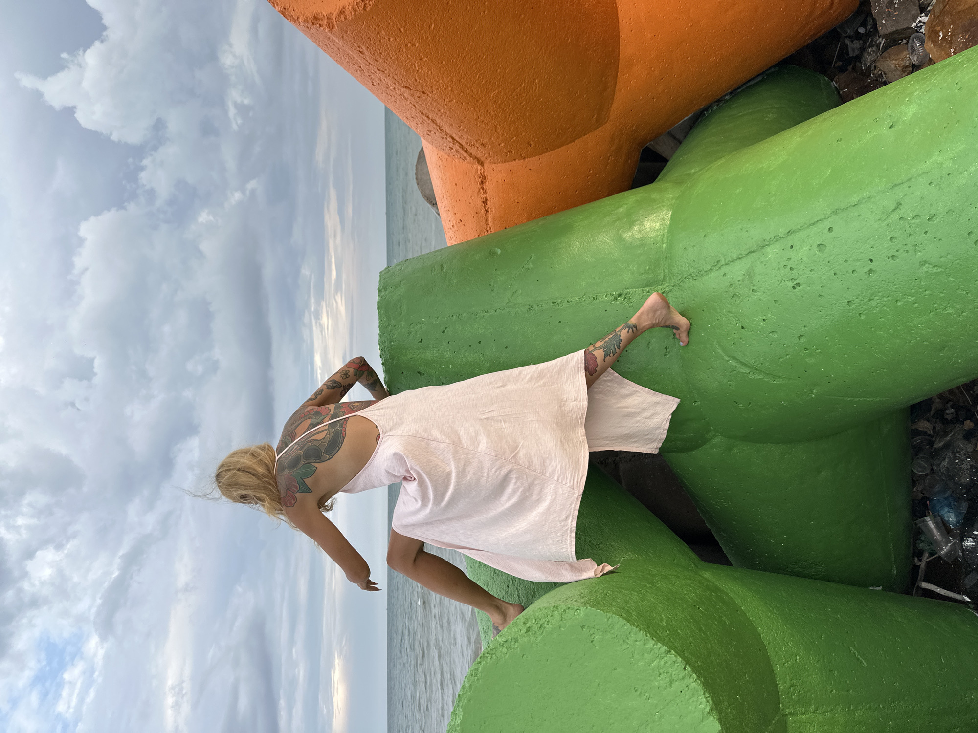 The image depicts a woman posing or moving gracefully on large, brightly colored concrete breakwaters at a coastal location. The woman, with blonde hair, is seen from behind, wearing a light pink, sleeveless dress and is barefoot. She has extensive, colorful tattoos visible on her arms, shoulders, and upper back. Her body is leaned forward, with one leg extended, as if she is balancing or in mid-stride, looking out towards the sea.
In the foreground and midground, massive concrete structures are painted in vibrant green and orange, serving as sea defenses or tetrapods. The calm sea is visible in the background, stretching to the horizon under an overcast sky with soft, diffused clouds. The lighting suggests it is daytime. Some debris, including plastic items, can be seen accumulated in the crevices between the concrete blocks. There is no visible text in the image. The setting suggests a coastal area, likely in Phu Thuy Ward, Vietnam, given the context.