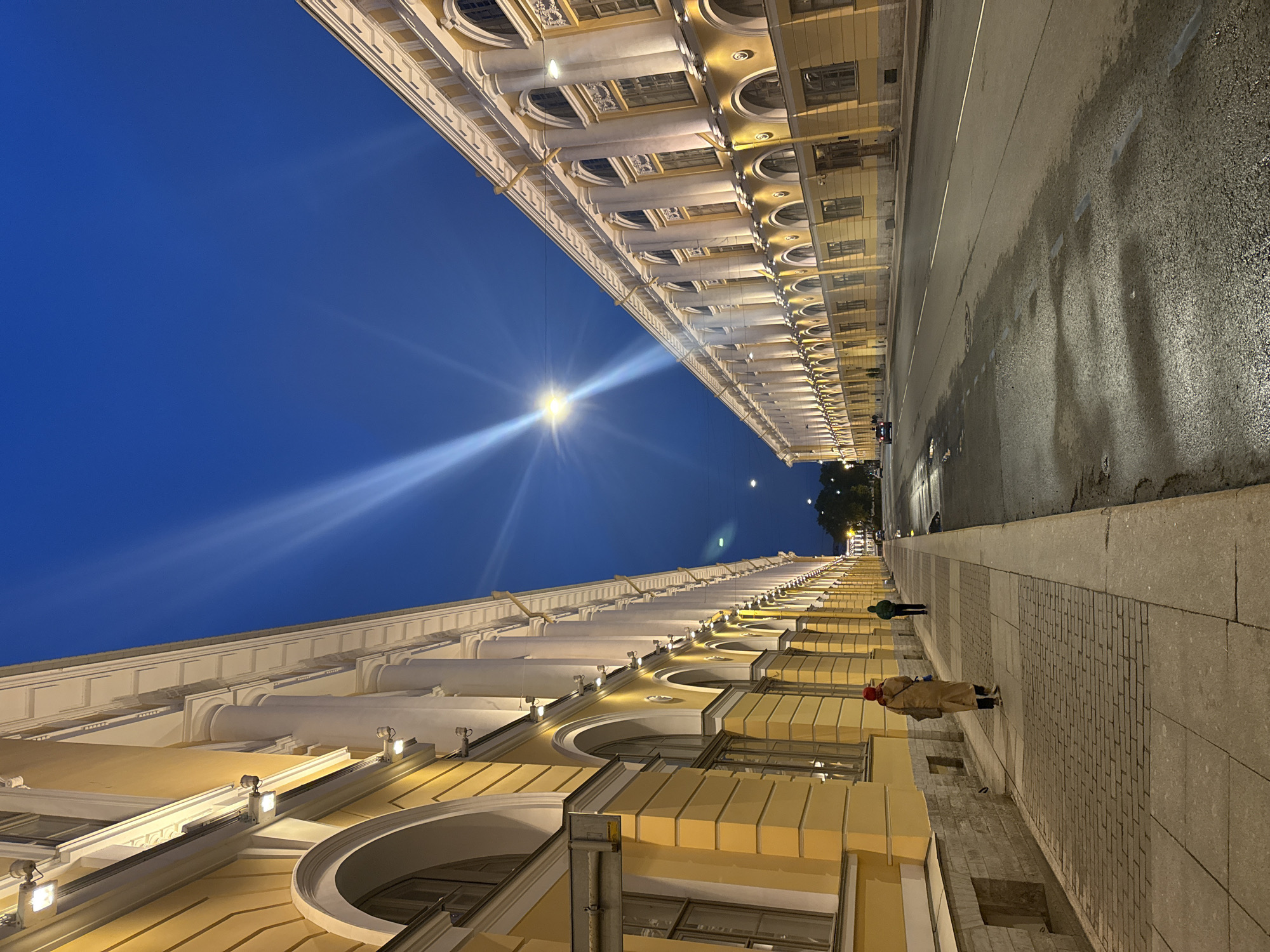 The image captures a street scene in Saint Petersburg, Russia, at dusk. On either side of the street are buildings of classical architecture with colonnades and arched windows. A street lamp shines brightly between the buildings. The street is wet, reflecting the city lights. On the sidewalk, two pedestrians are walking, one wearing a tan coat and a red hat, and the other wearing a green coat.