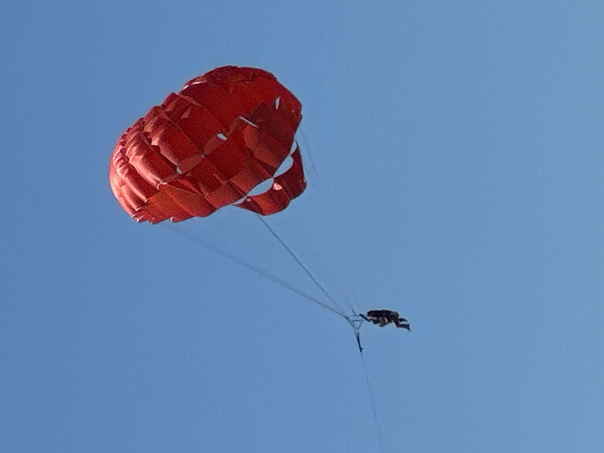 A person is parasailing high in the sky above Budva, Montenegro. The parasail is bright red and billows out, catching the wind. The person is suspended below in a harness, with their legs dangling. The sky is a clear, vibrant blue.