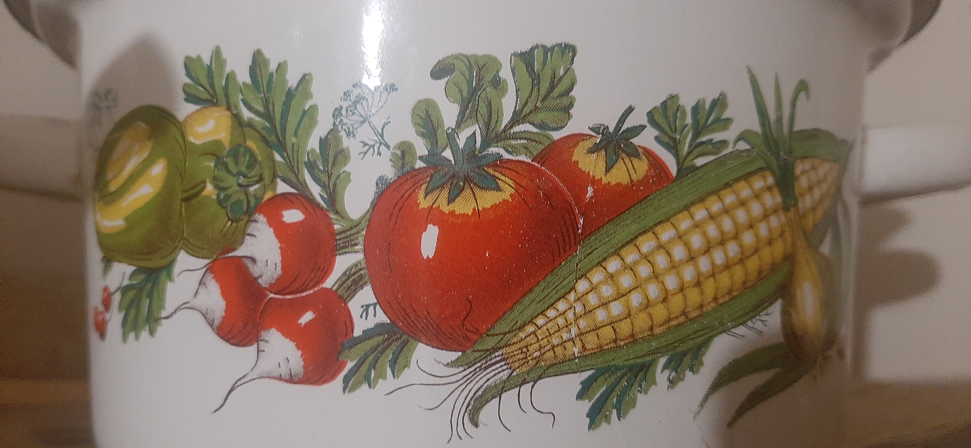 A close-up, eye-level shot shows a section of a white enamel pot decorated with a colorful illustration of fresh produce. The artwork features two bright red tomatoes with yellow tops, a bundle of radishes with white bulbs and red skin, a green pepper with yellow accents, and a cob of corn with yellow kernels and green husks. Lush green leaves and delicate dill sprigs are scattered throughout the arrangement. A hint of a handle is visible on the right side of the pot. The background is out of focus, appearing as a plain, light-colored surface. The scene is static and lacks any environmental context that would suggest a specific location in Yessentukskaya, Russia, or any other city.