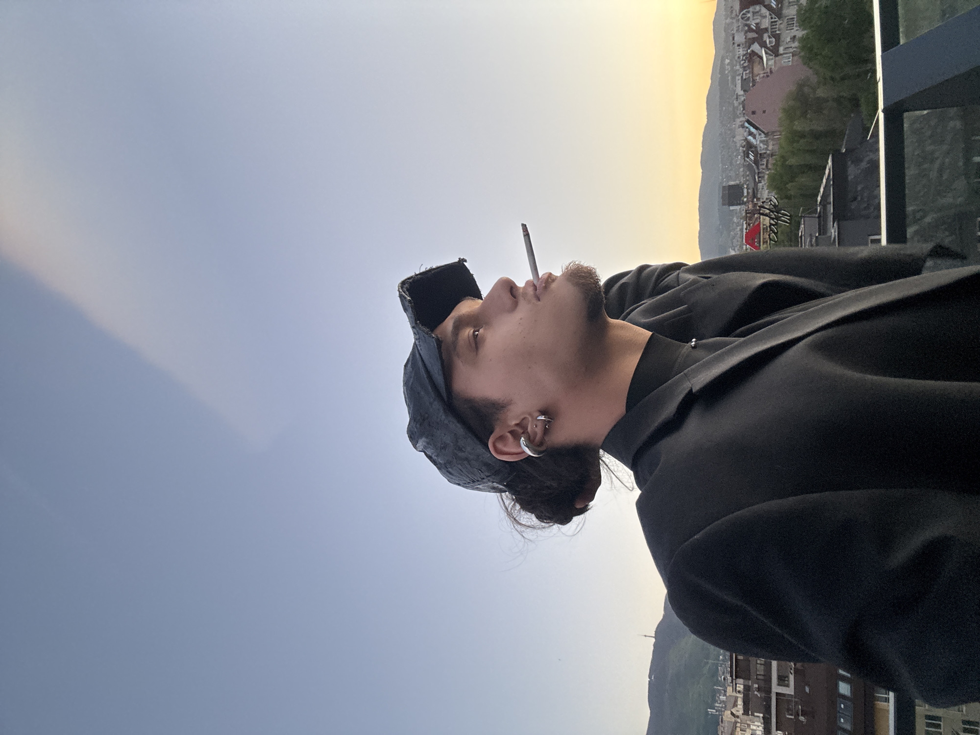 A man with an earring and a stubbled beard is smoking a cigarette. He is wearing a black jacket and a hat. The backdrop consists of a cityscape transitioning into a horizon with a sunset. The location cannot be determined as Sofia, Bulgaria.