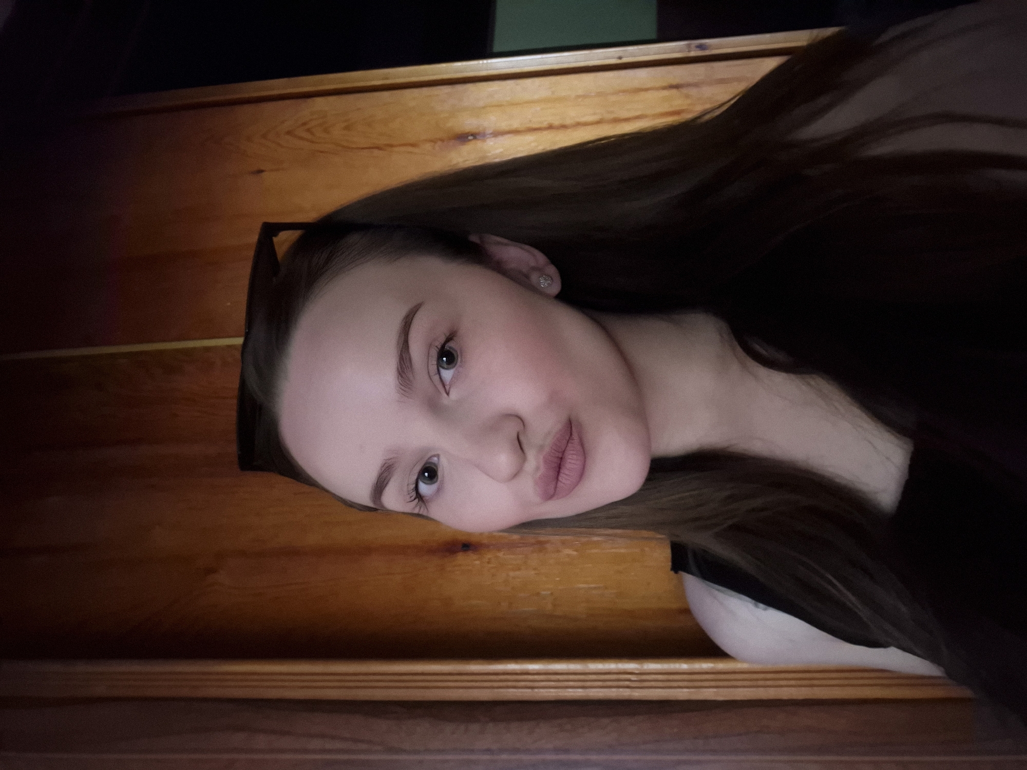 A young Caucasian woman with light skin, long brown hair, and a black top is pictured from the chest up, taking a selfie. She is looking directly at the camera with her lips pursed, and her eyebrows are arched. She is wearing a small, sparkly stud earring in her left ear. Sunglasses are perched on top of her head. The background is a wooden door with vertical planks, and to the right of the door, a dark space is visible. The lighting in the image is dim.