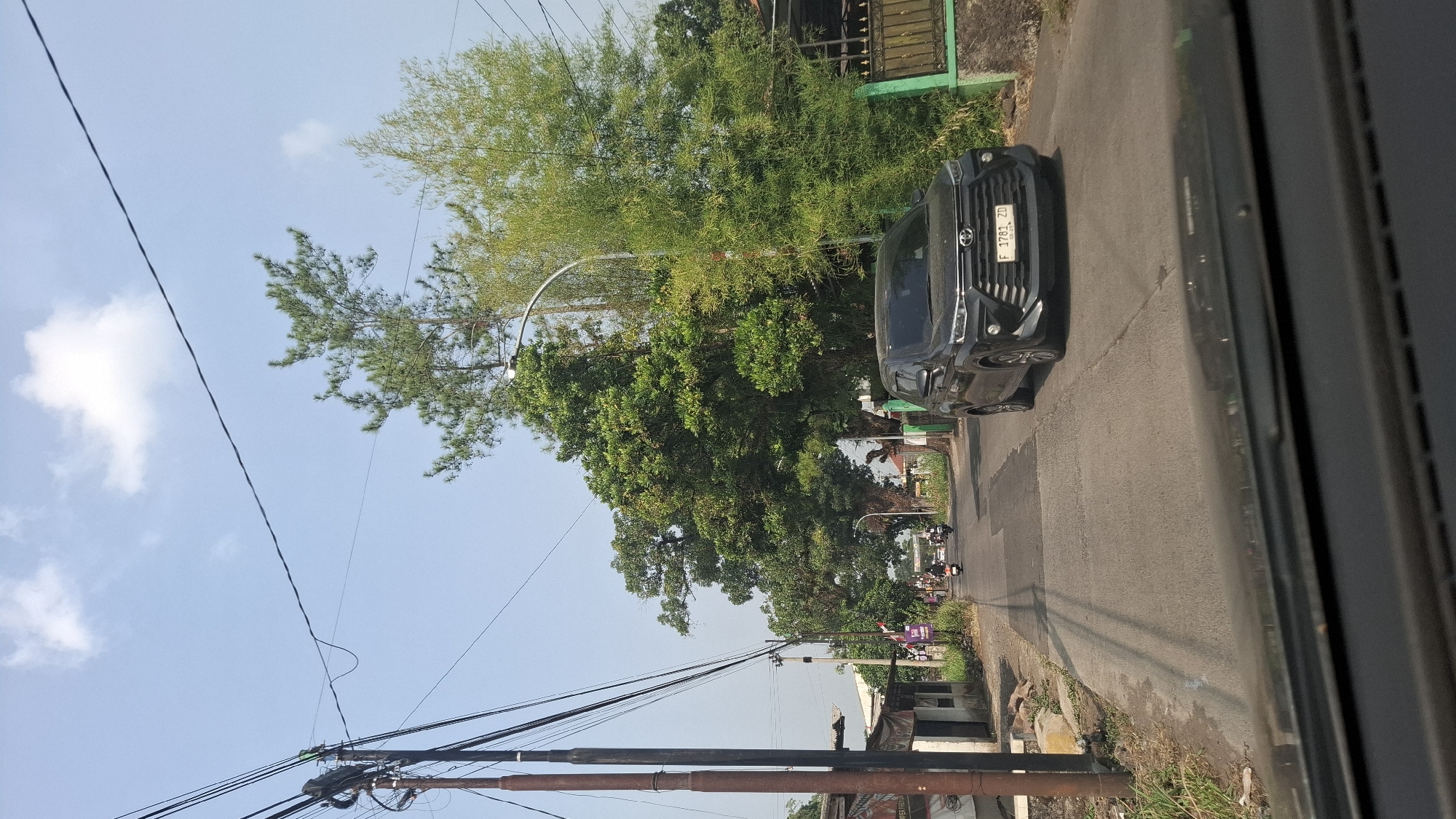 The image shows a street scene in Cilaku, Indonesia. A black Toyota Avanza is driving on the right side of the road. In the background, there are several houses and trees. The sky is blue with some white clouds. The road appears to be a two-lane street with some patches of repair. There are power lines running overhead, supported by a wooden utility pole on the left side of the image. A few motorcycles are visible further down the road, suggesting traffic. A green metal fence is visible on the right side of the road, adjacent to a house. The overall environment appears to be a residential area.