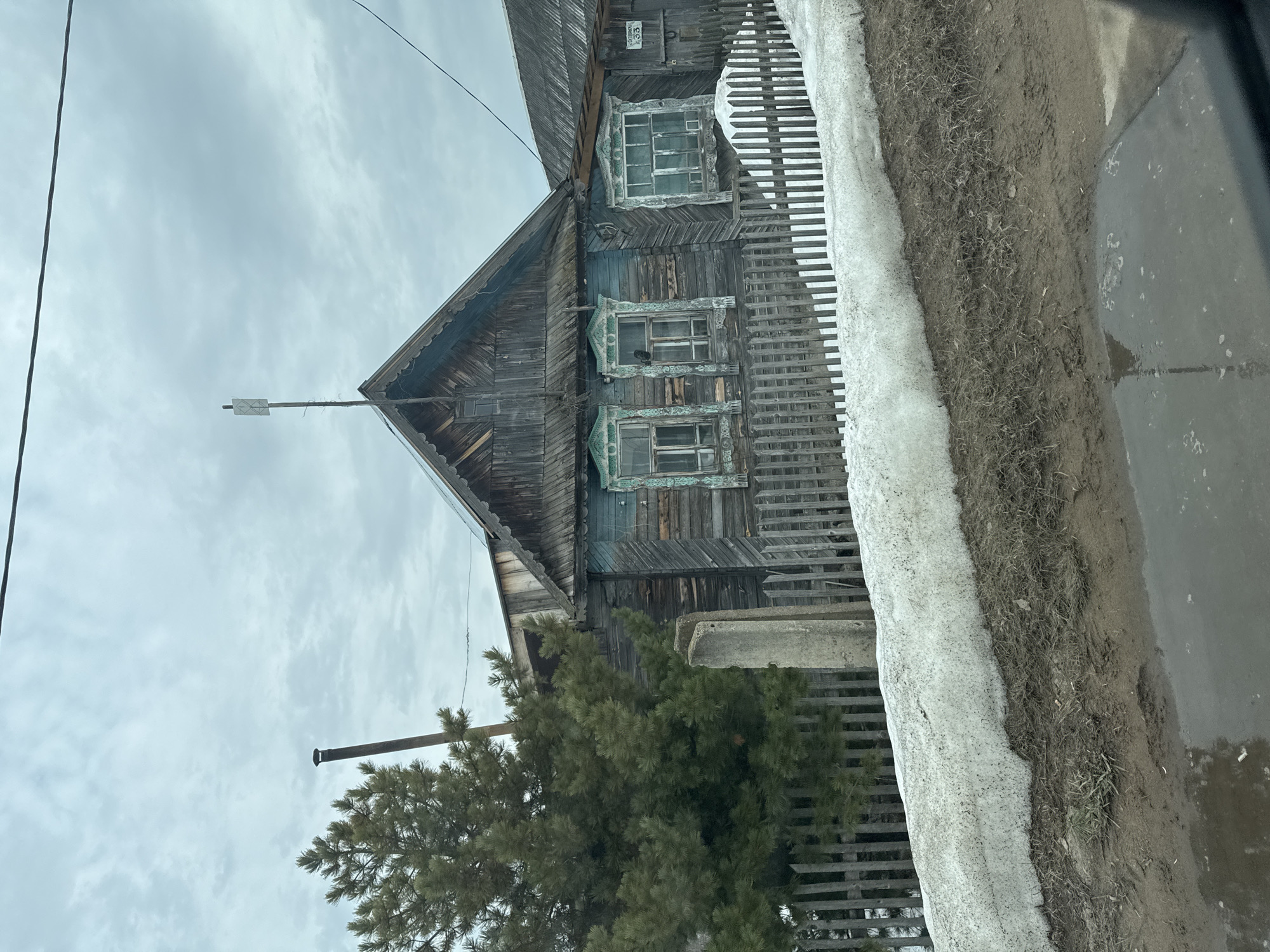 The image shows a two-story wooden house in Noviy, Russia. The house is weathered, with faded blue paint and intricate wood trim around the windows. It has a steeply pitched roof with a metal antenna on top. A wooden fence partially surrounds the property. There is snow along the side of the road with a muddy section separating it from a puddle of standing water. A pine tree is visible in the front yard. The sky is overcast. The number "33" is visible on the house. A utility pole with a chimney is also visible in the lower left.