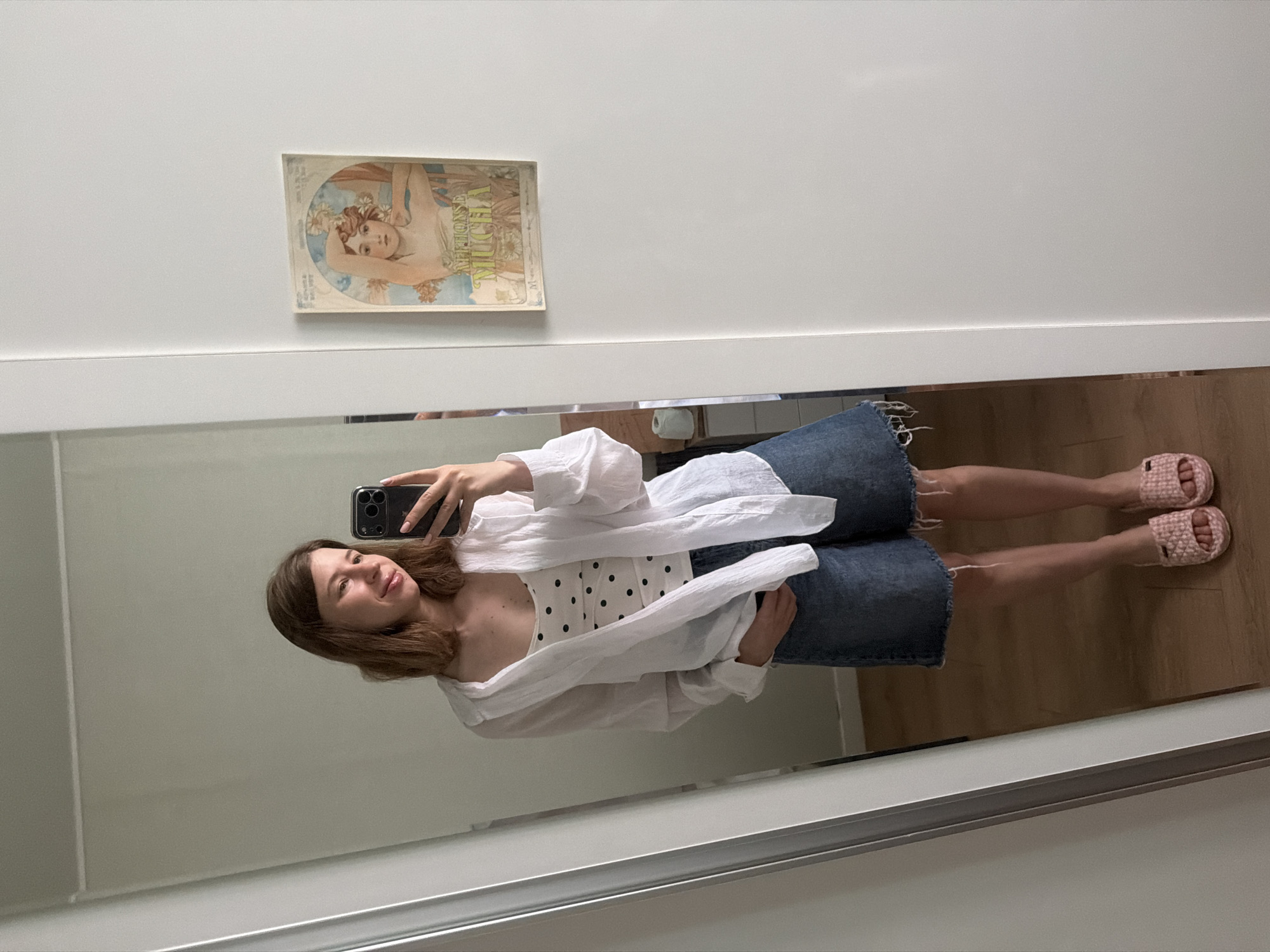 A young woman with medium-length brown hair is taking a mirror selfie in a brightly lit room. She is wearing a white polka-dot tank top layered under an open white linen shirt, denim shorts with a frayed hem, and pink slip-on sandals. She is holding a black smartphone in her right hand, and her left hand is casually tucked into the pocket of her shorts. The background shows a large mirror reflecting the woman, a glimpse of a door frame, and a framed Art Nouveau print on the wall. The floor appears to be made of light-colored wood. The overall impression is of a casual, domestic setting, possibly a bedroom or dressing area, in Dongducheon-si, South Korea.