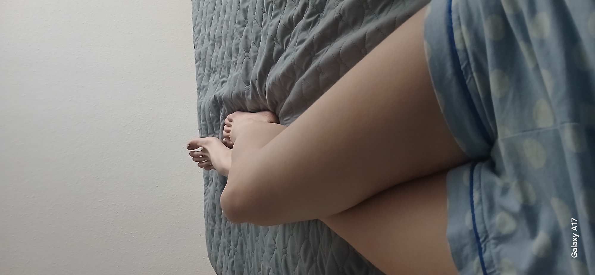 A close-up shot captures a person lying on a gray quilted bedspread. Only their legs are visible, with one leg crossed over the other. The individual is wearing blue and white polka-dot pajamas. A white wall is visible in the background to the left of the bed. A "Galaxy A17" watermark is visible on the bottom right corner of the image. No specific location in Yekaterinburg, Russia, is identifiable from the image.